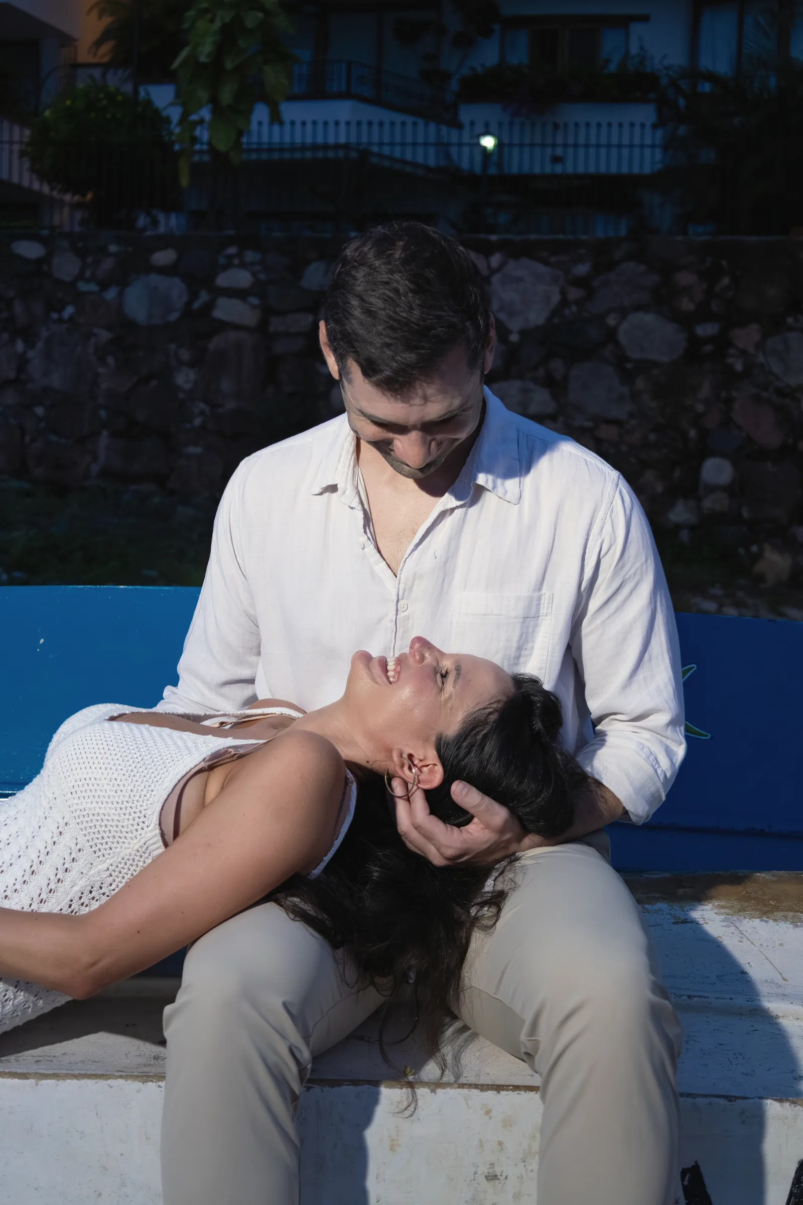 Carmela leaning back into Charlie's lap as he looks down at her, seated together on a blue boat at dusk