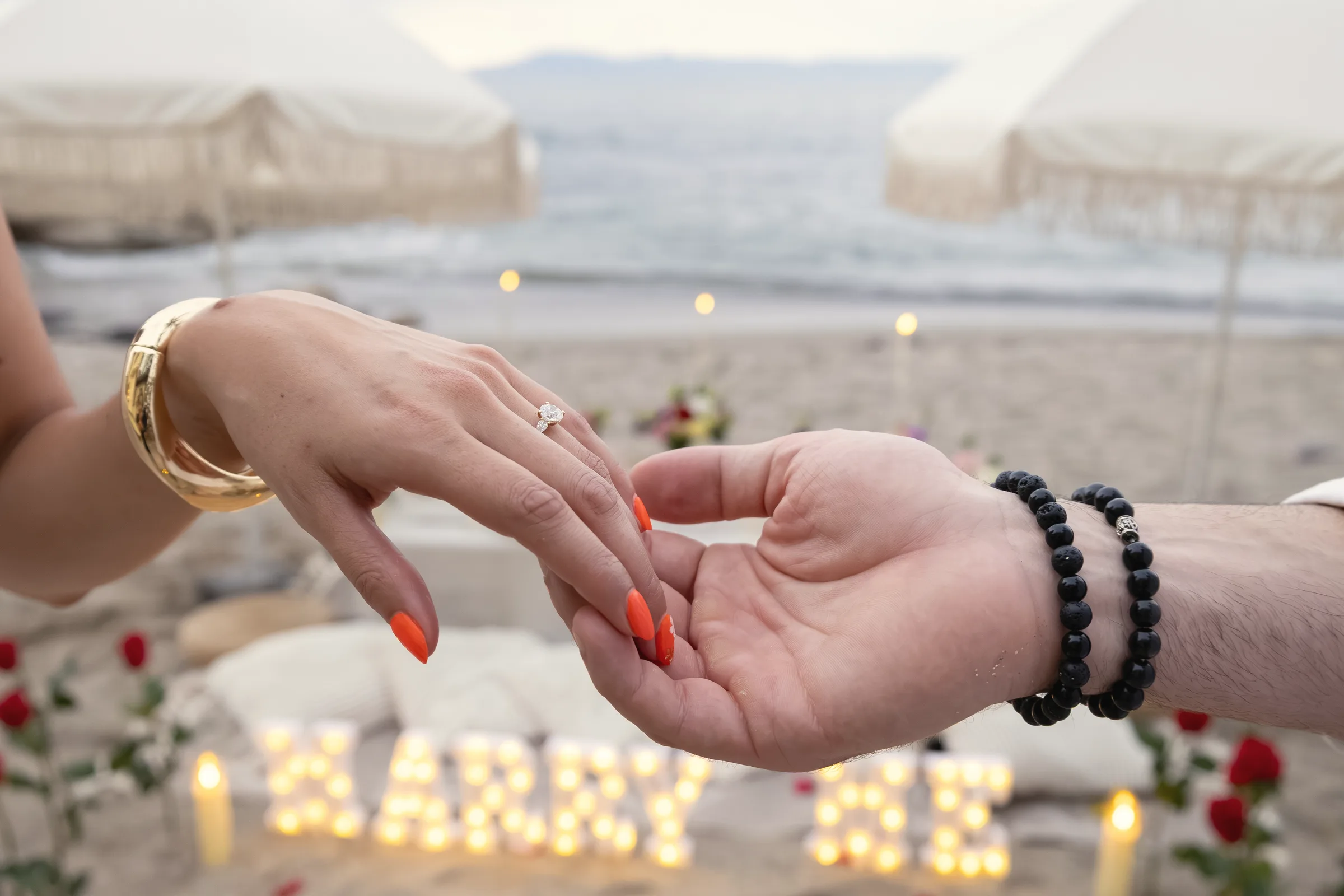 Carmela's hand with her engagement ring resting in Charlie's hand, a lit MARRY ME sign and rose petals on the sand behind