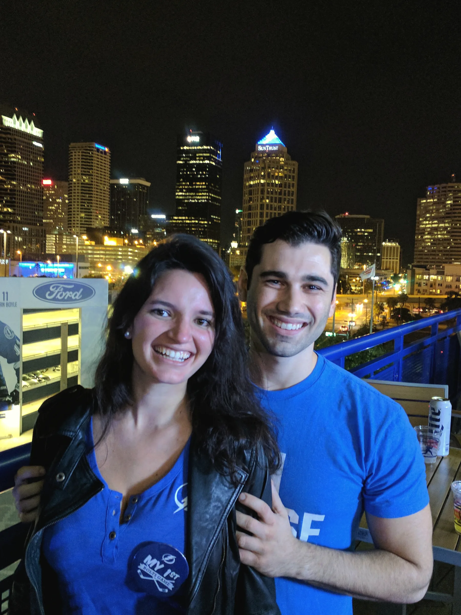 Carmela and Charlie smiling in a close selfie on a balcony at night, a lit city skyline behind them