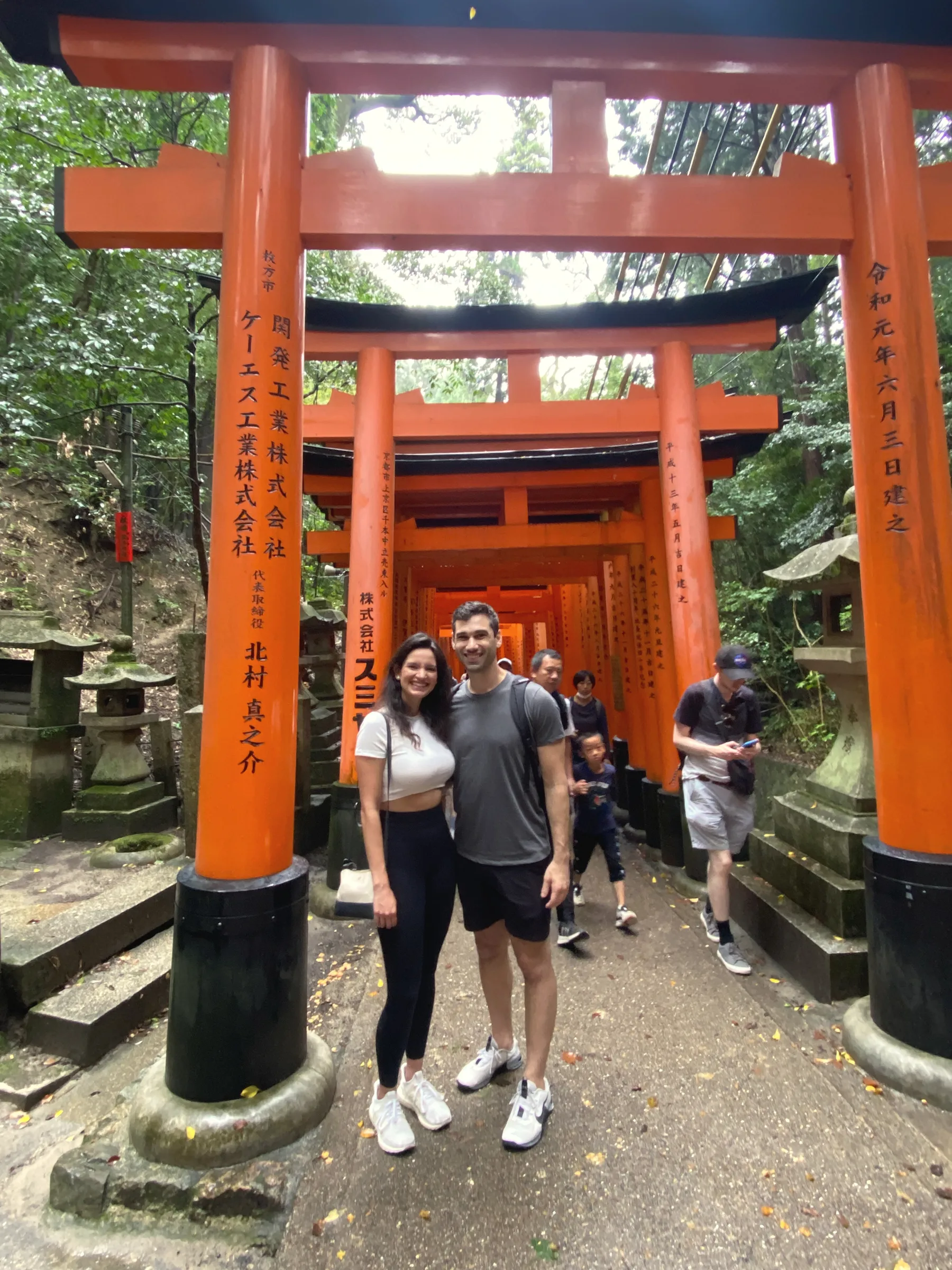 Carmela and Charlie standing on a path between rows of tall vermilion torii gates in a forest