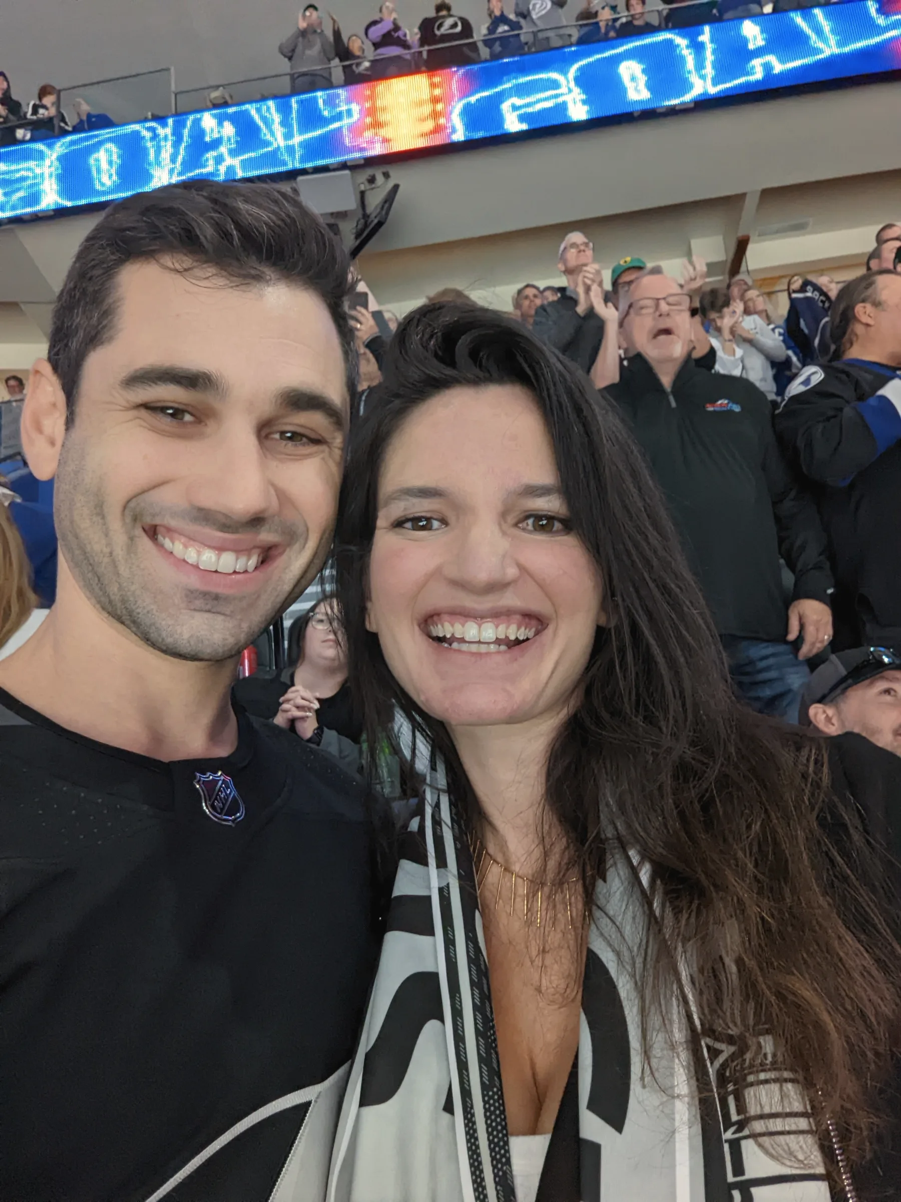 Carmela and Charlie smiling close to the camera in a crowded arena, a glowing scoreboard above them