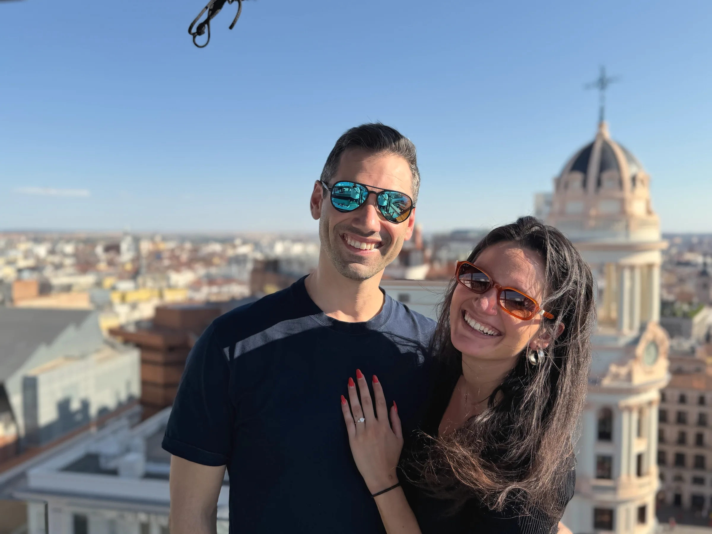 Carmela and Charlie in sunglasses on a rooftop, her hand on his chest, with a domed building and city rooftops behind them