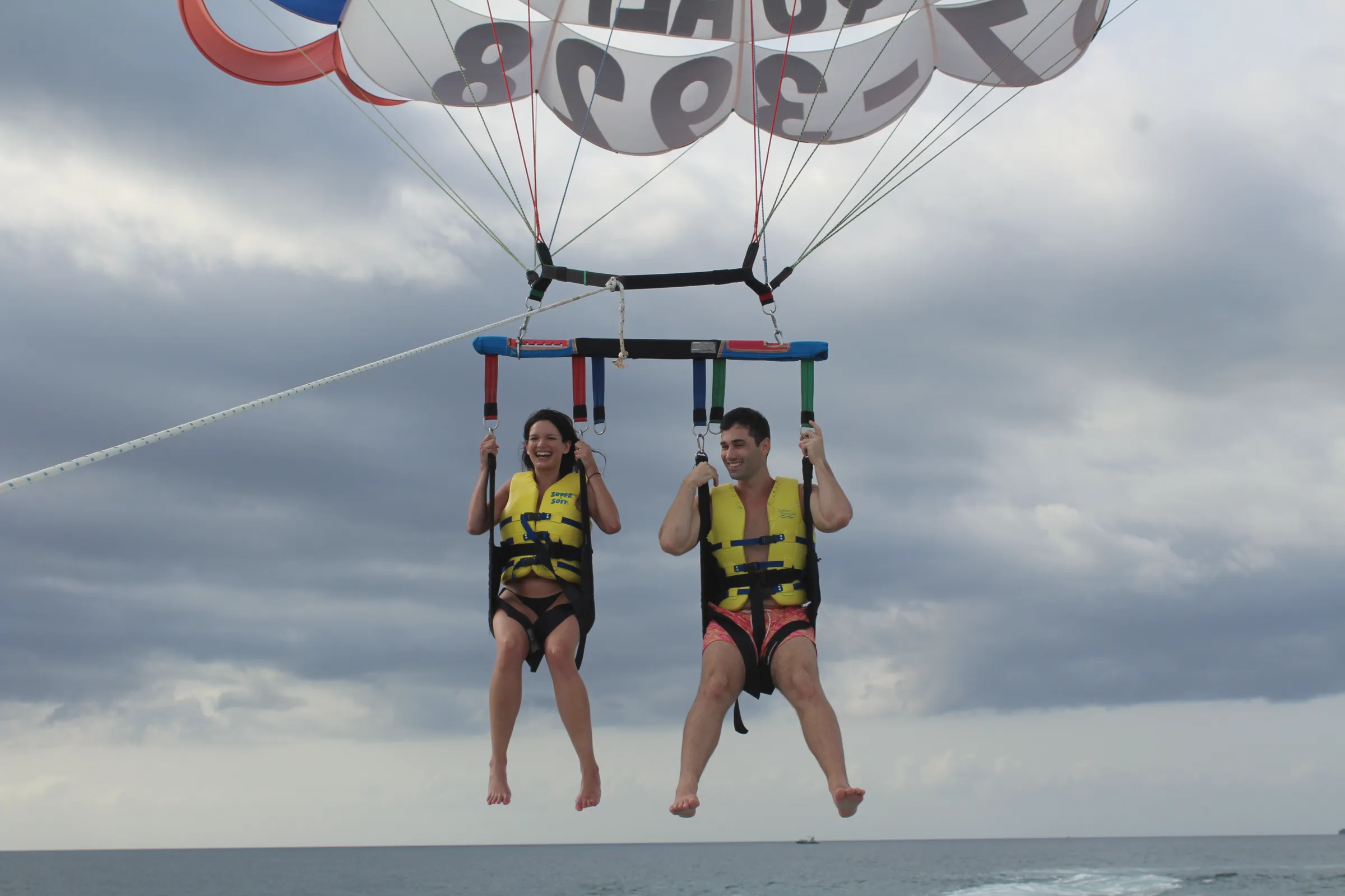 Carmela and Charlie side by side in yellow life vests, dangling beneath a parachute over the sea