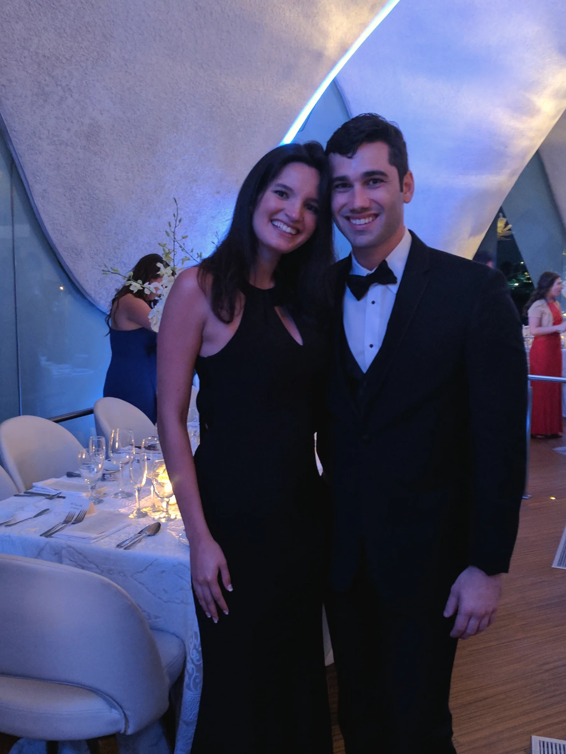 Carmela in a black dress and Charlie in a tuxedo with a bow tie, in front of a set banquet table at an evening event