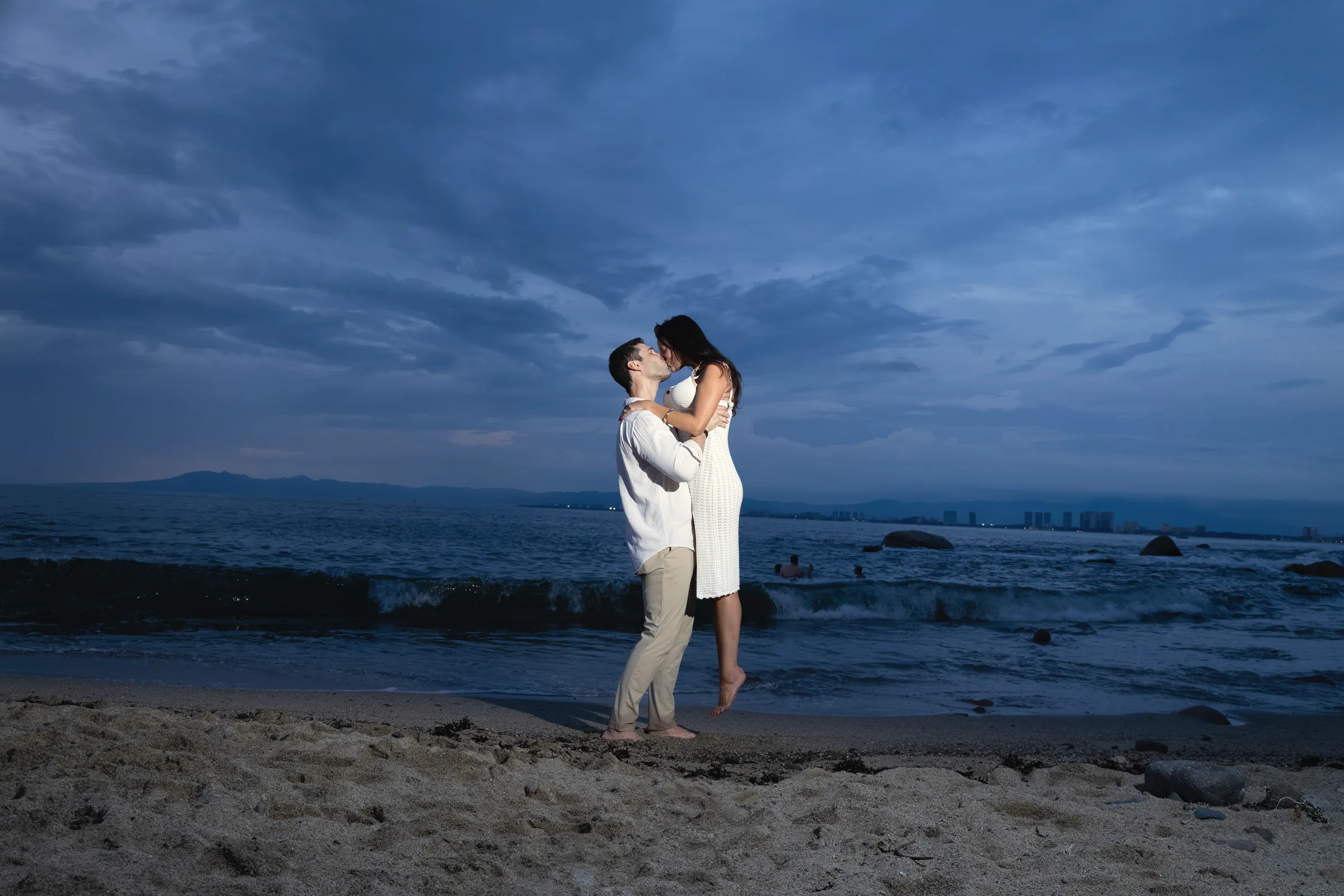 Carmela and Charlie embracing small on the sand under a deep blue evening sky, the ocean behind them