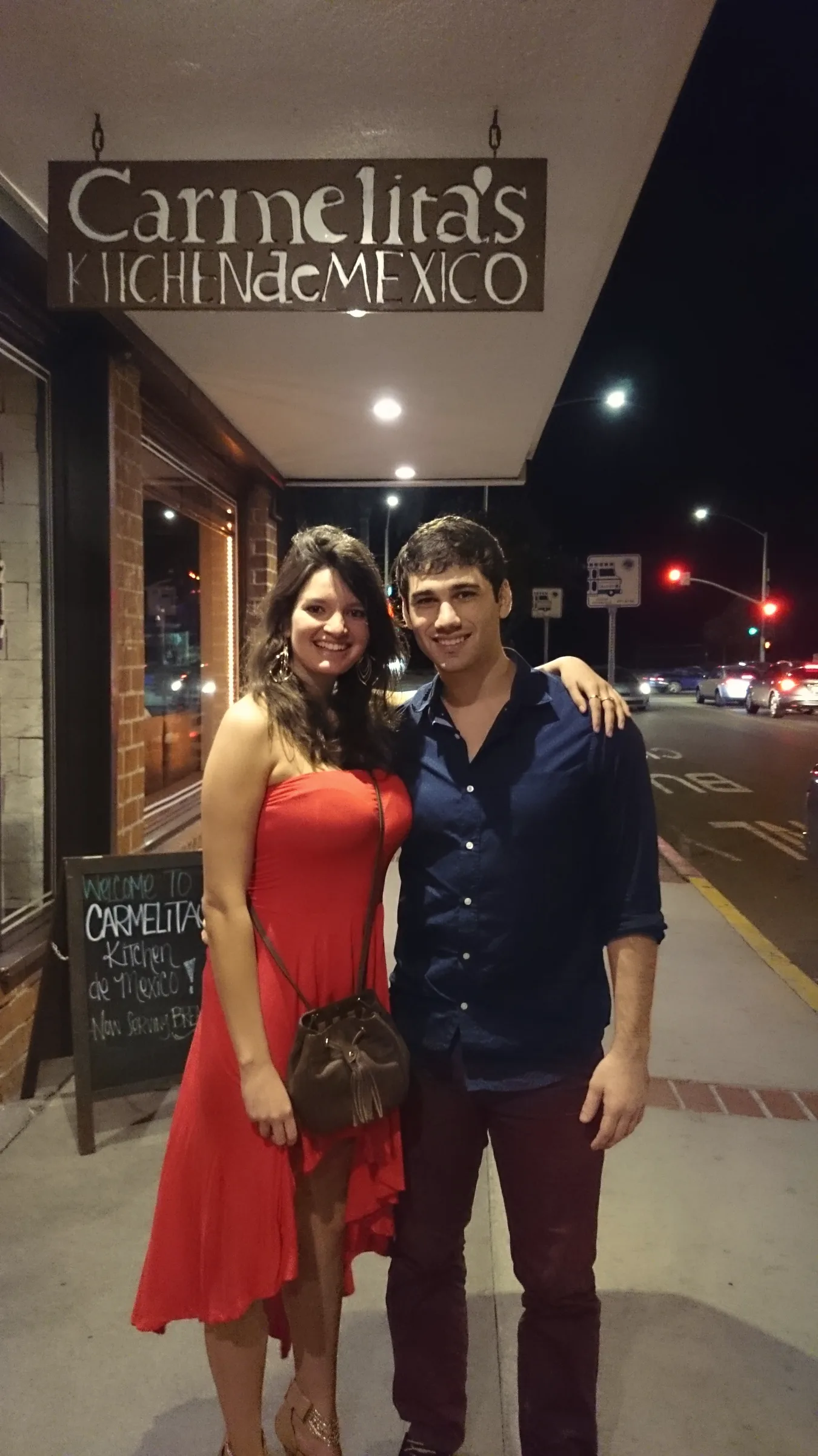 Carmela in a red dress and Charlie in a navy shirt with their arms around each other on a street at night, in front of a restaurant sign reading Carmelita's Kitchen de Mexico