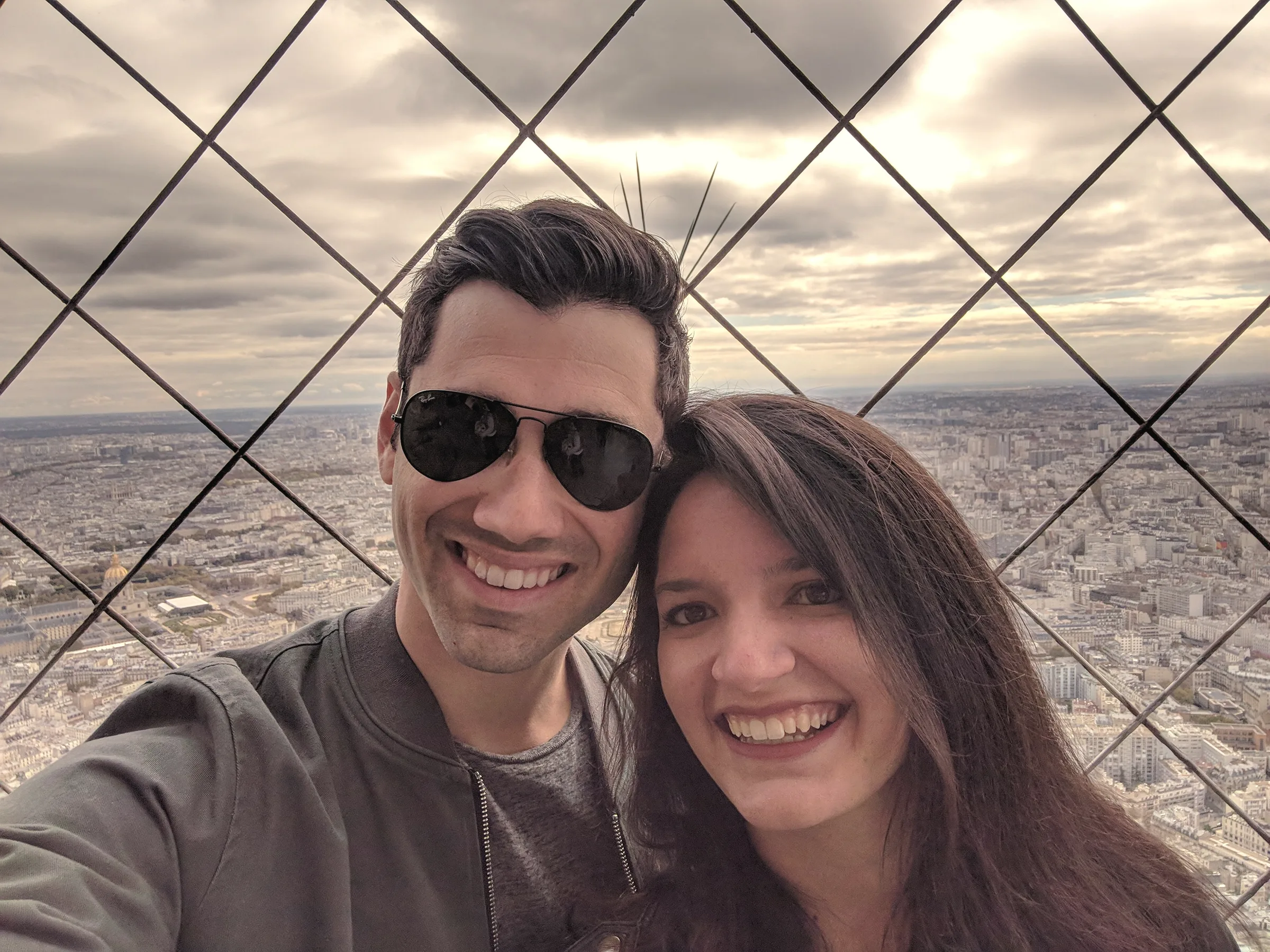 Carmela and Charlie in a close selfie behind a lattice of steel bars, a wide city and cloudy sky below