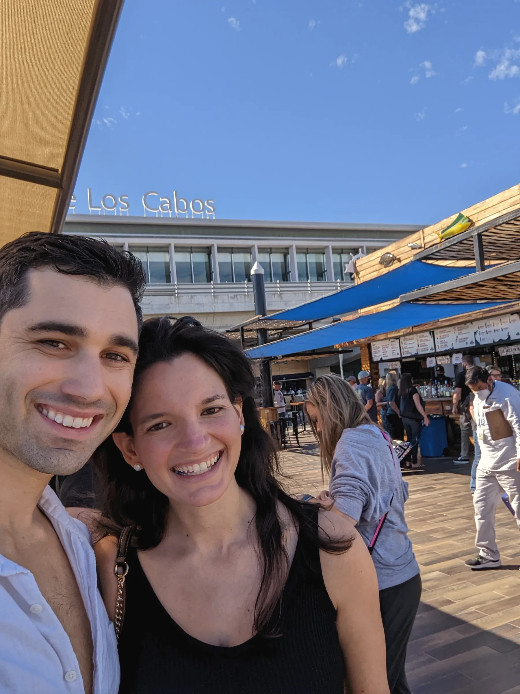 Carmela and Charlie leaning together, smiling, in front of a waterfront building with a Los Cabos sign and blue shade sails