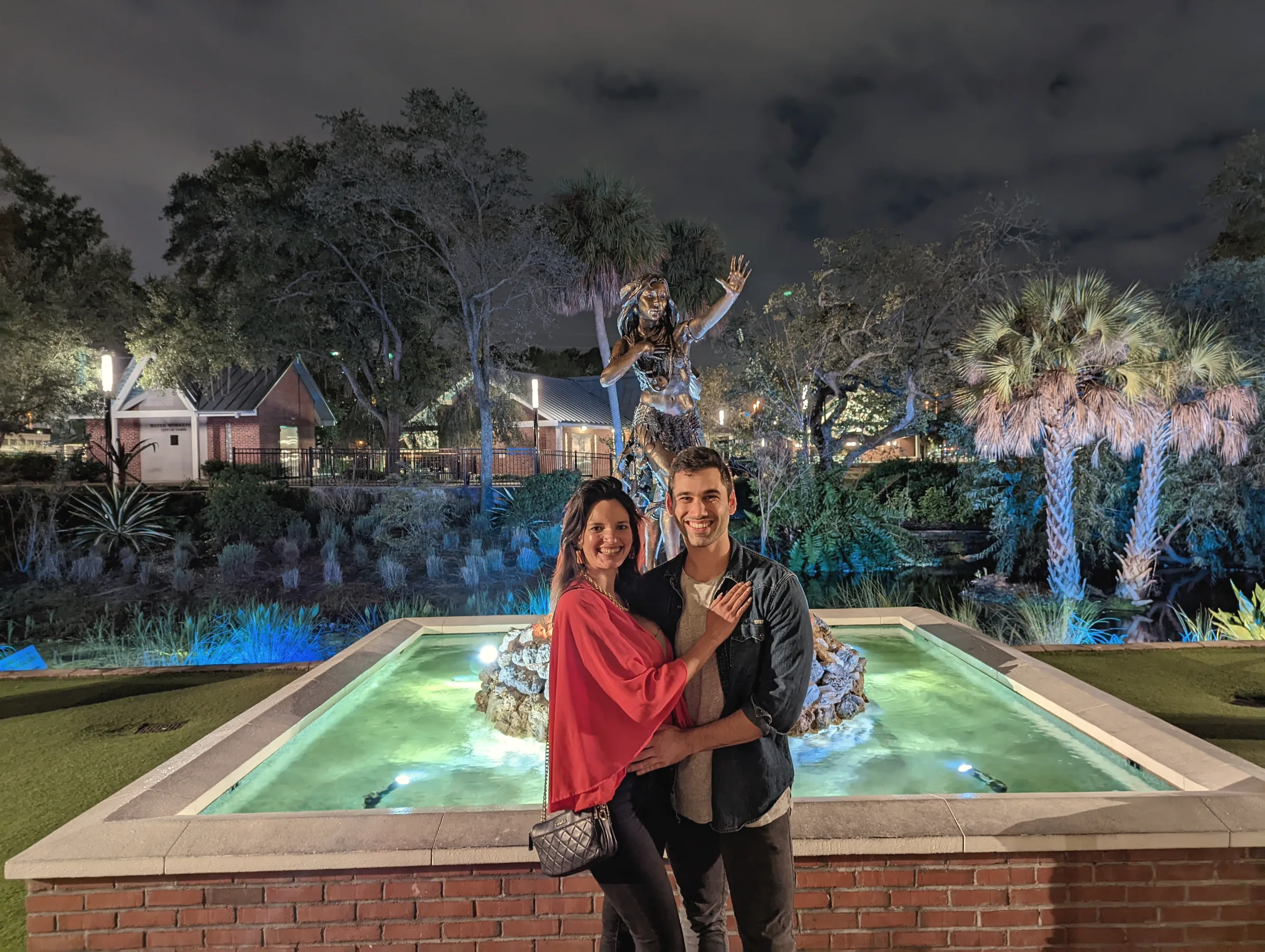 Carmela in red and Charlie in a dark jacket in front of an illuminated fountain and palm trees at night