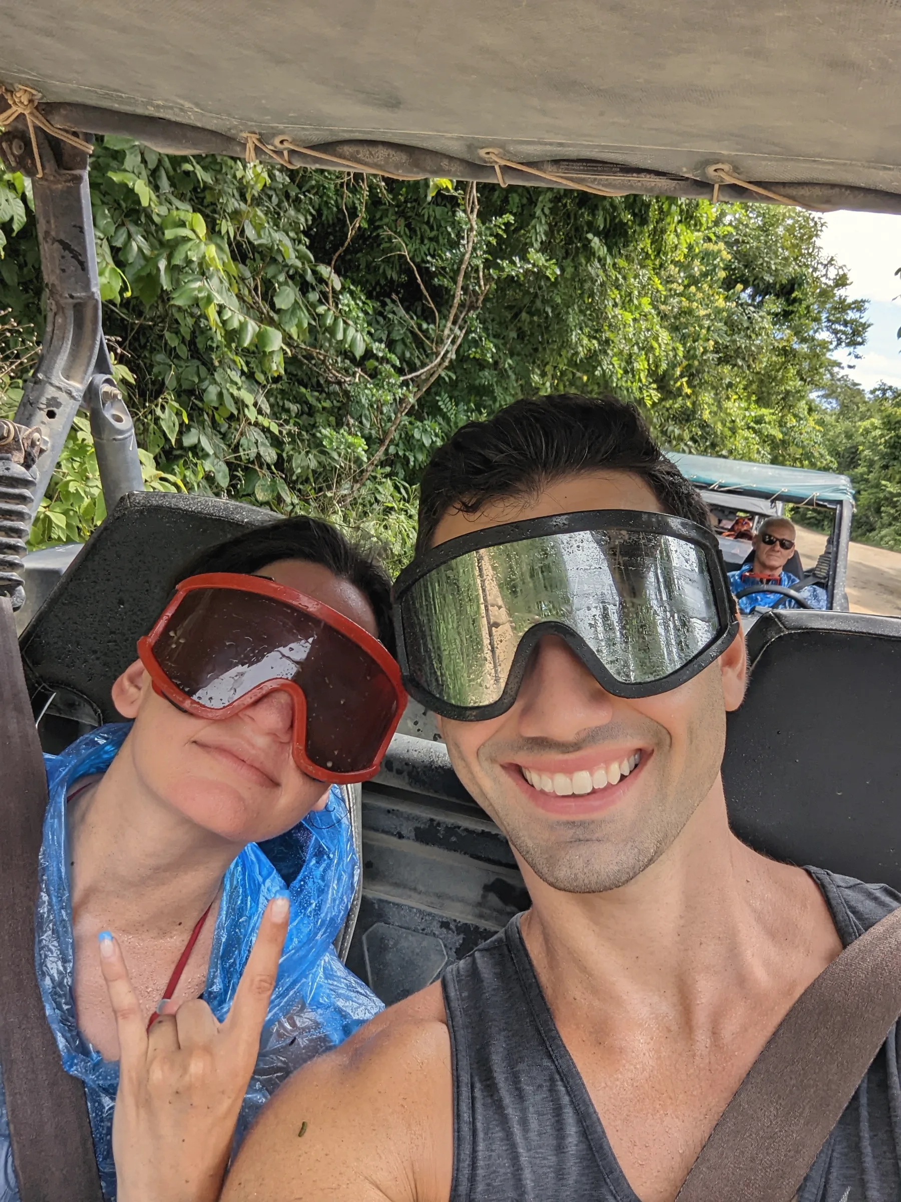 Carmela in red goggles and a blue poncho beside Charlie in goggles, both grinning in an open off-road vehicle with jungle behind
