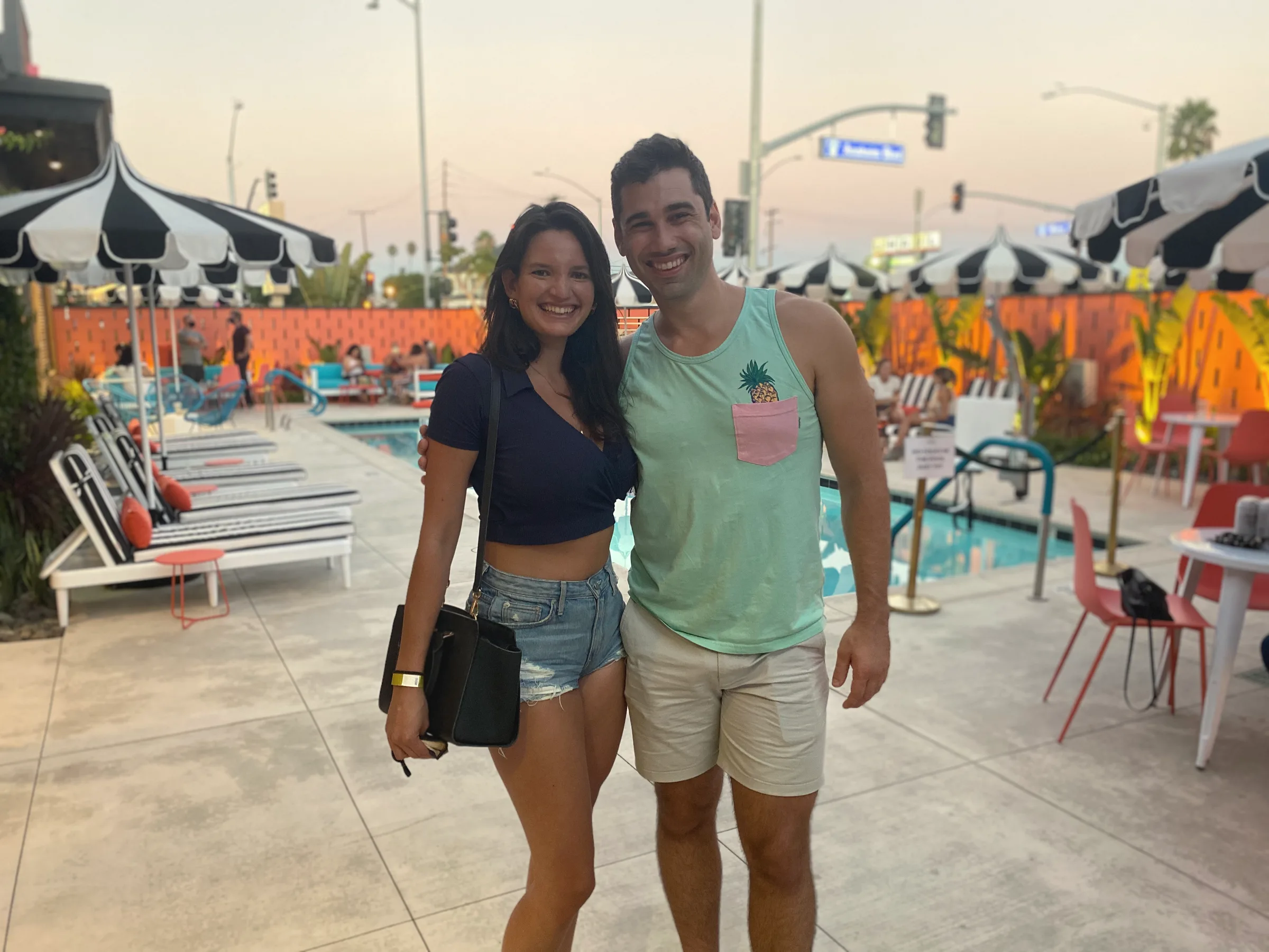 Carmela and Charlie standing side by side on a sunny pool deck with striped umbrellas and a palm-lined street