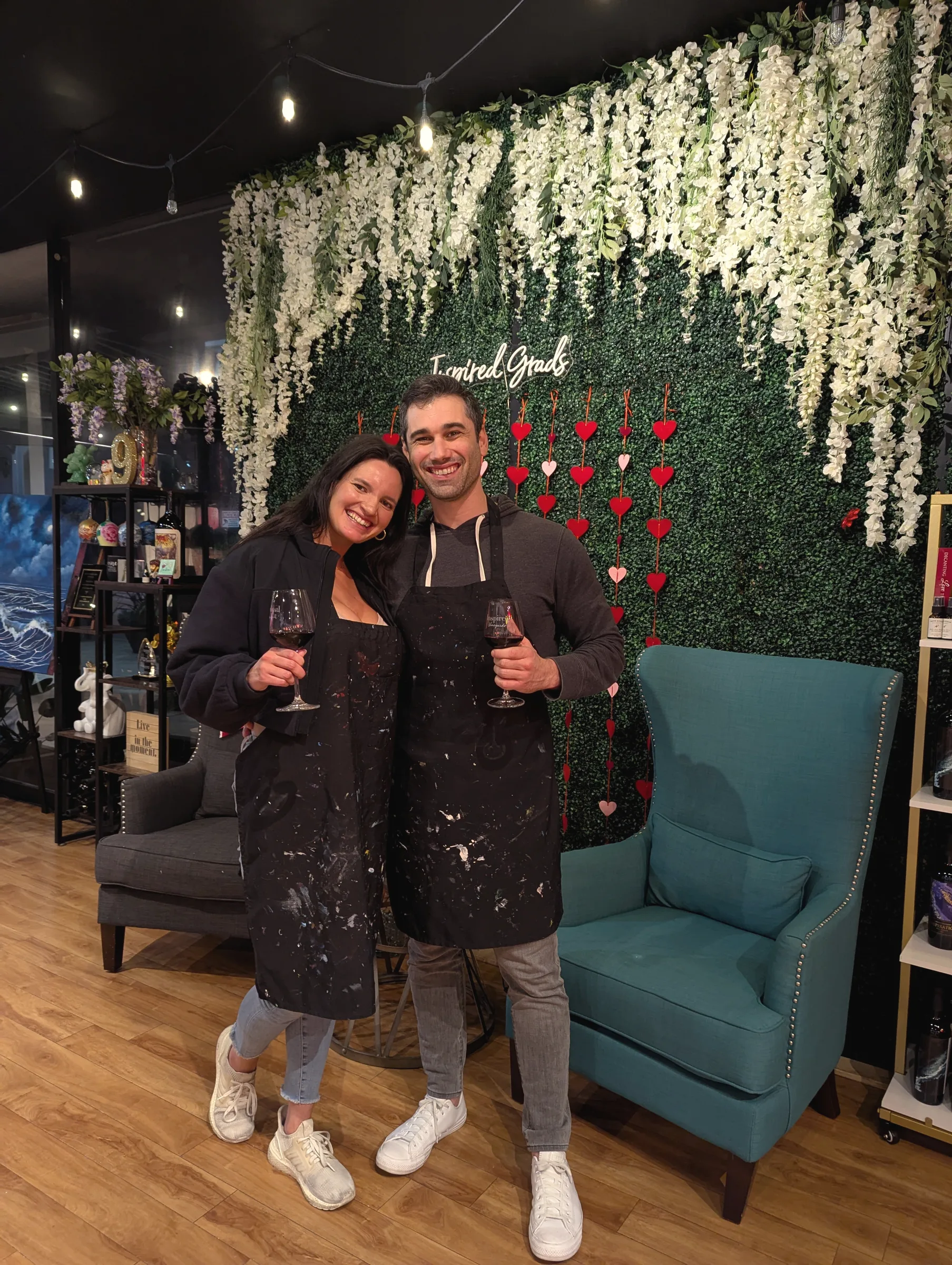 Carmela and Charlie in paint-splattered aprons, each holding a glass of wine, in front of a wall of white flowers