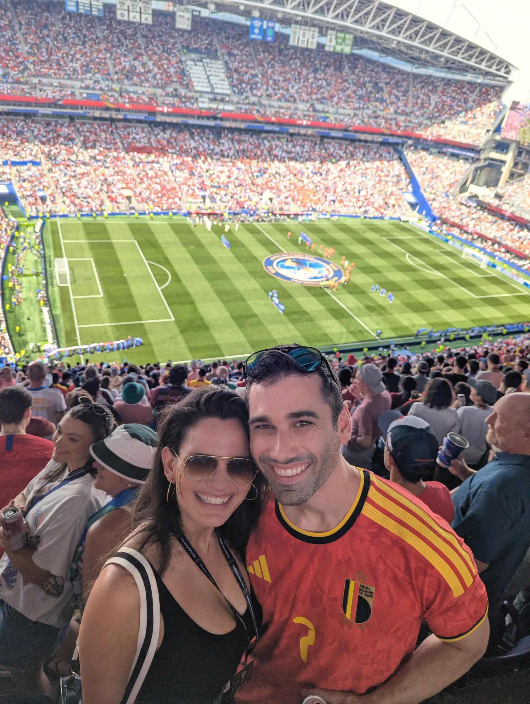 Carmela and Charlie in sunglasses, cheek to cheek in the stands, a green pitch and packed stadium below