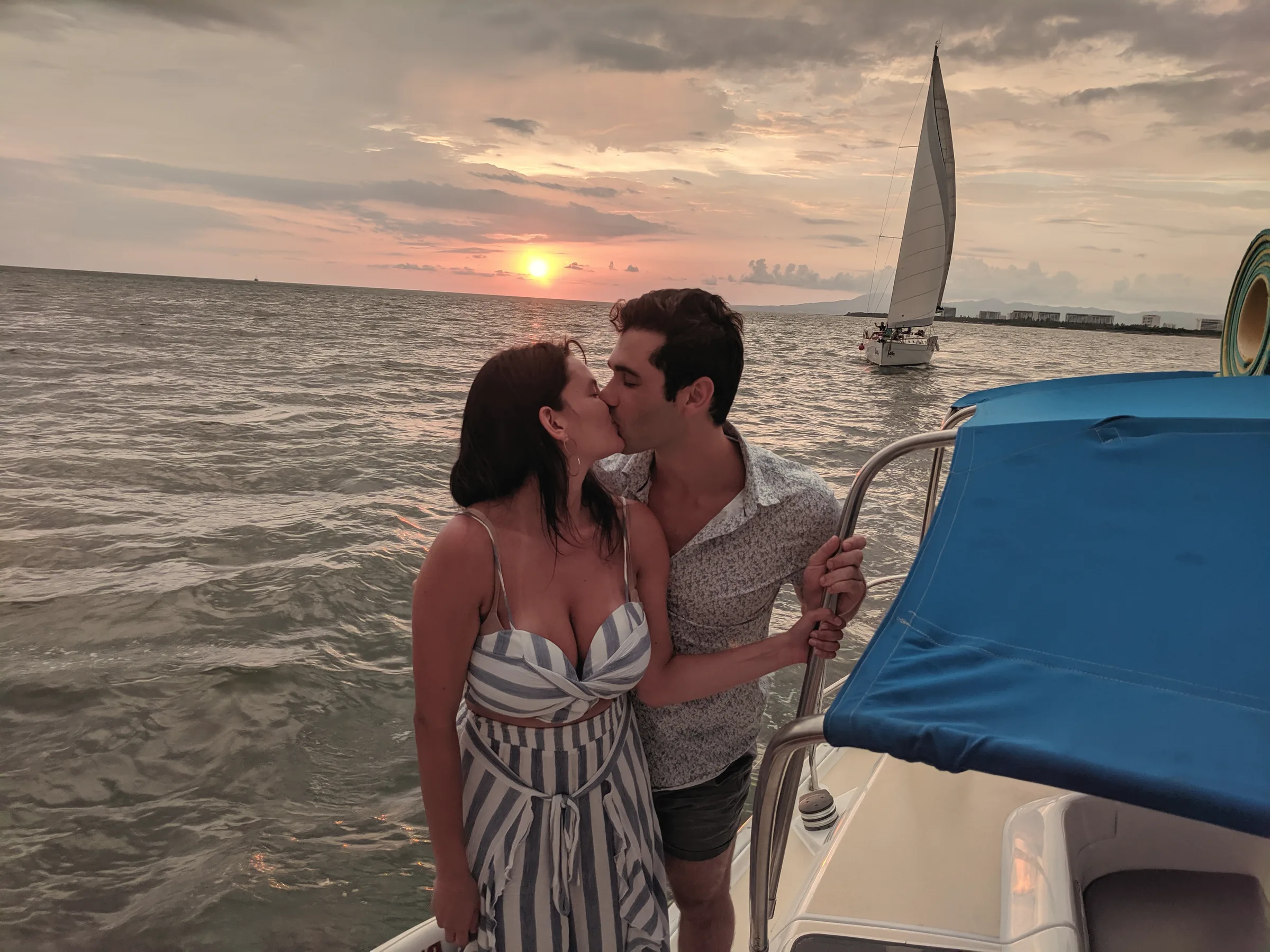 Carmela and Charlie kissing on the deck of a boat, a sailboat and the setting sun over the water behind them