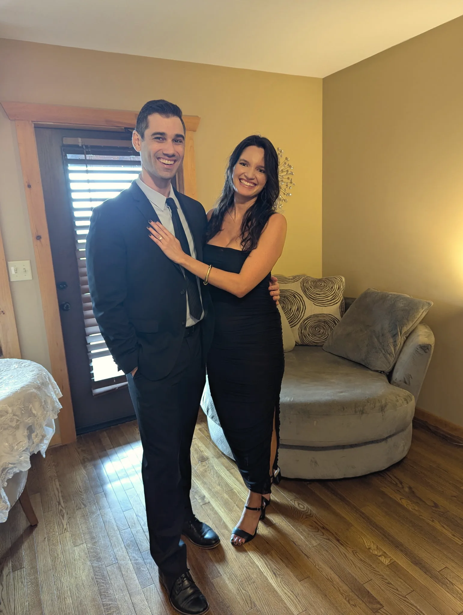 Charlie in a dark suit and tie and Carmela in a black dress, arm in arm in a warm wood-floored room