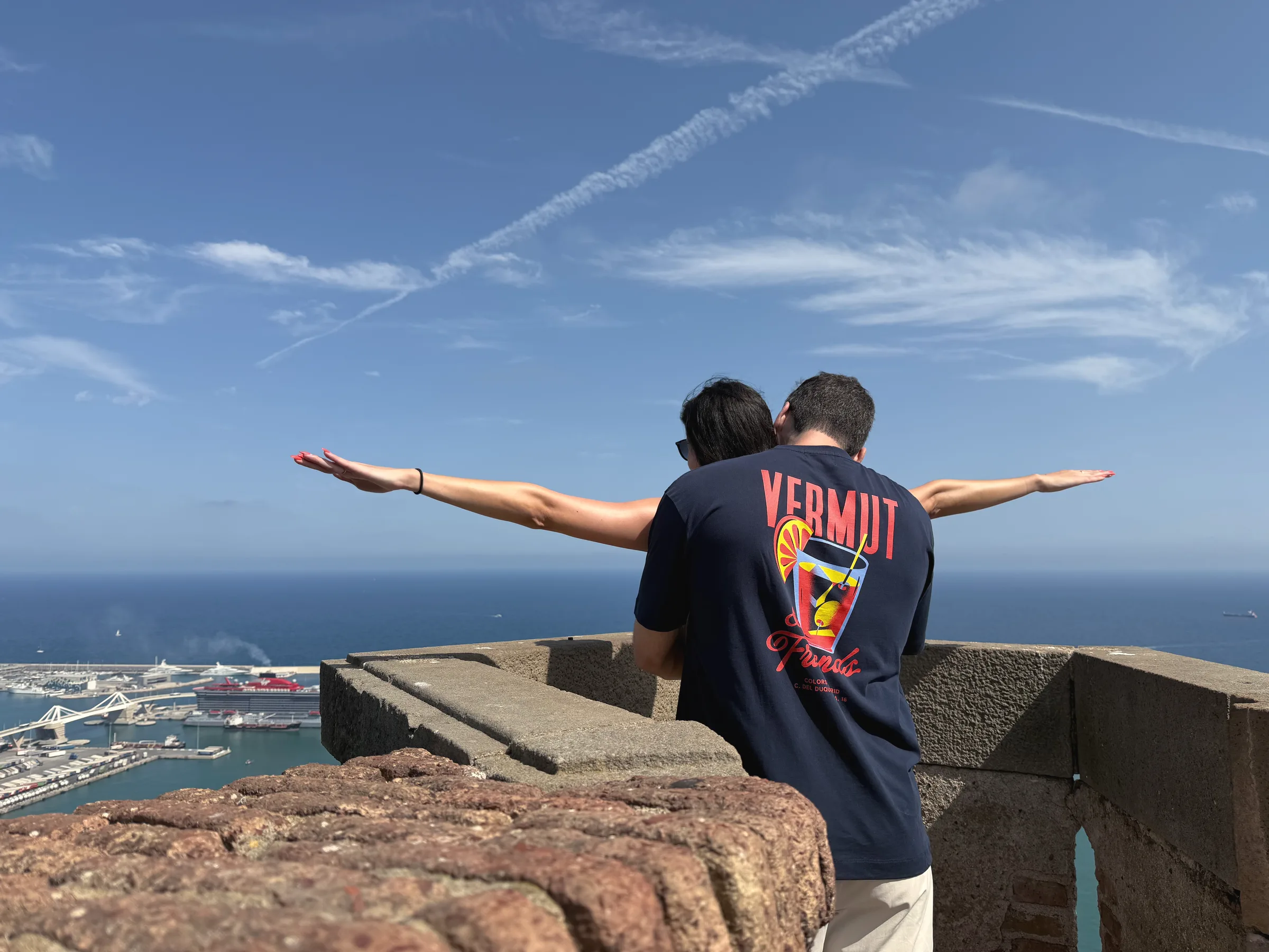 Carmela and Charlie seen from behind at a stone lookout, Carmela's arms stretched wide, a harbour and the sea far below