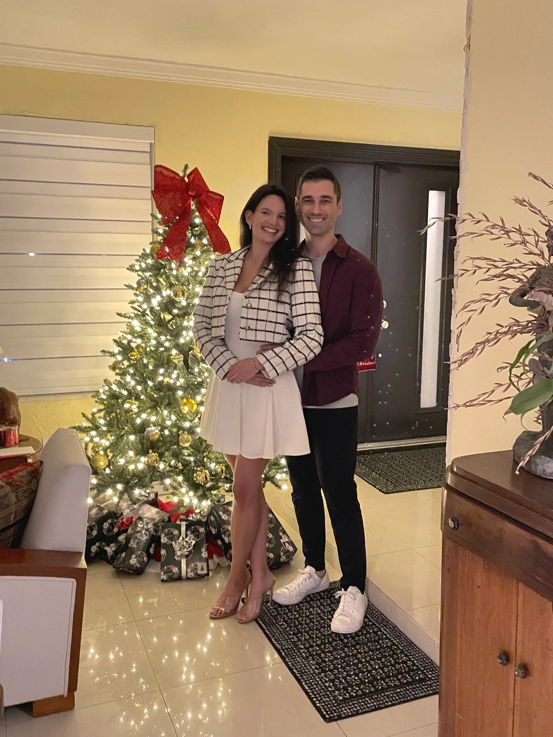 Carmela and Charlie arm in arm in a doorway beside a decorated Christmas tree with a big red bow