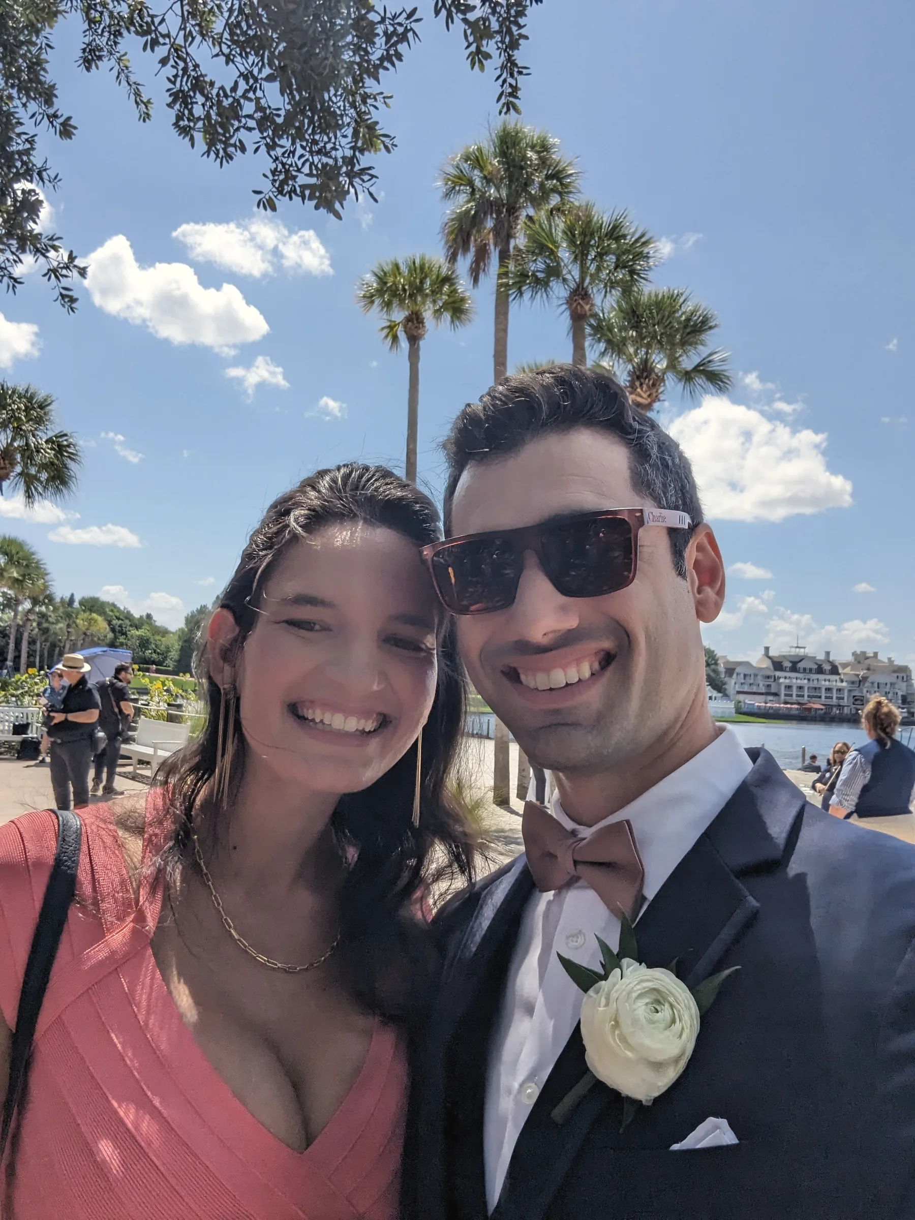Carmela in a coral dress and Charlie in a navy suit with a bow tie and white rose boutonniere, smiling under palm trees