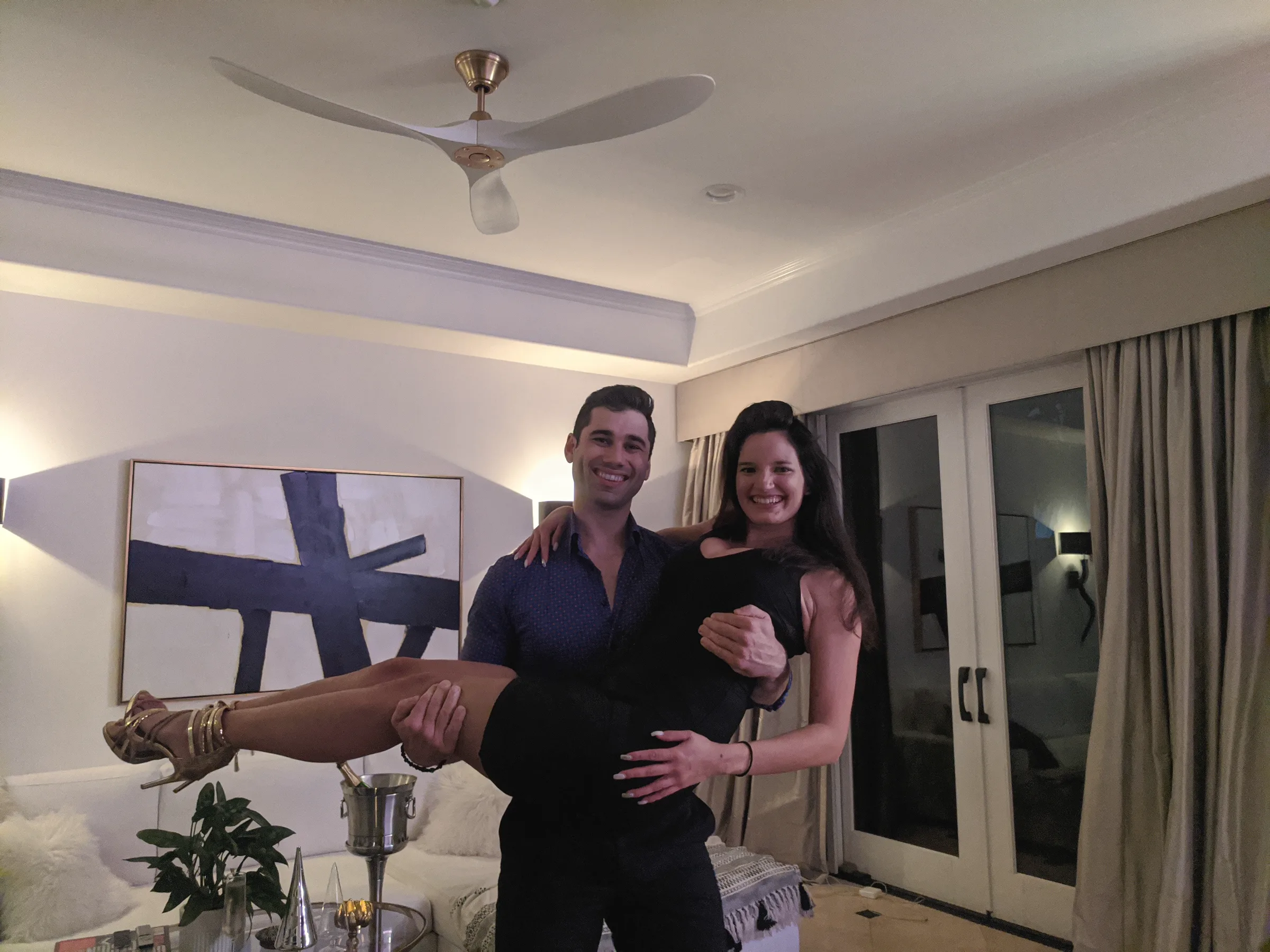 Charlie lifting Carmela off the ground in a bright room with a ceiling fan and french doors