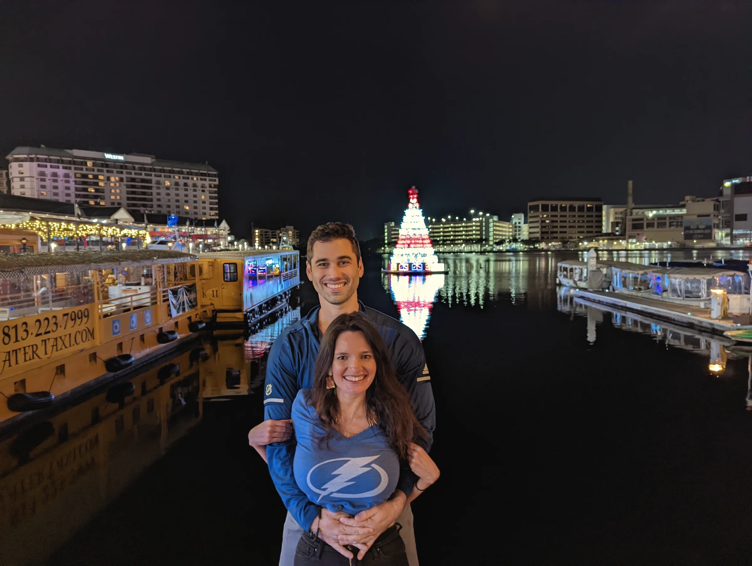 Carmela and Charlie in blue team shirts on a dark waterfront, boats and a lit Christmas tree reflected in the water behind