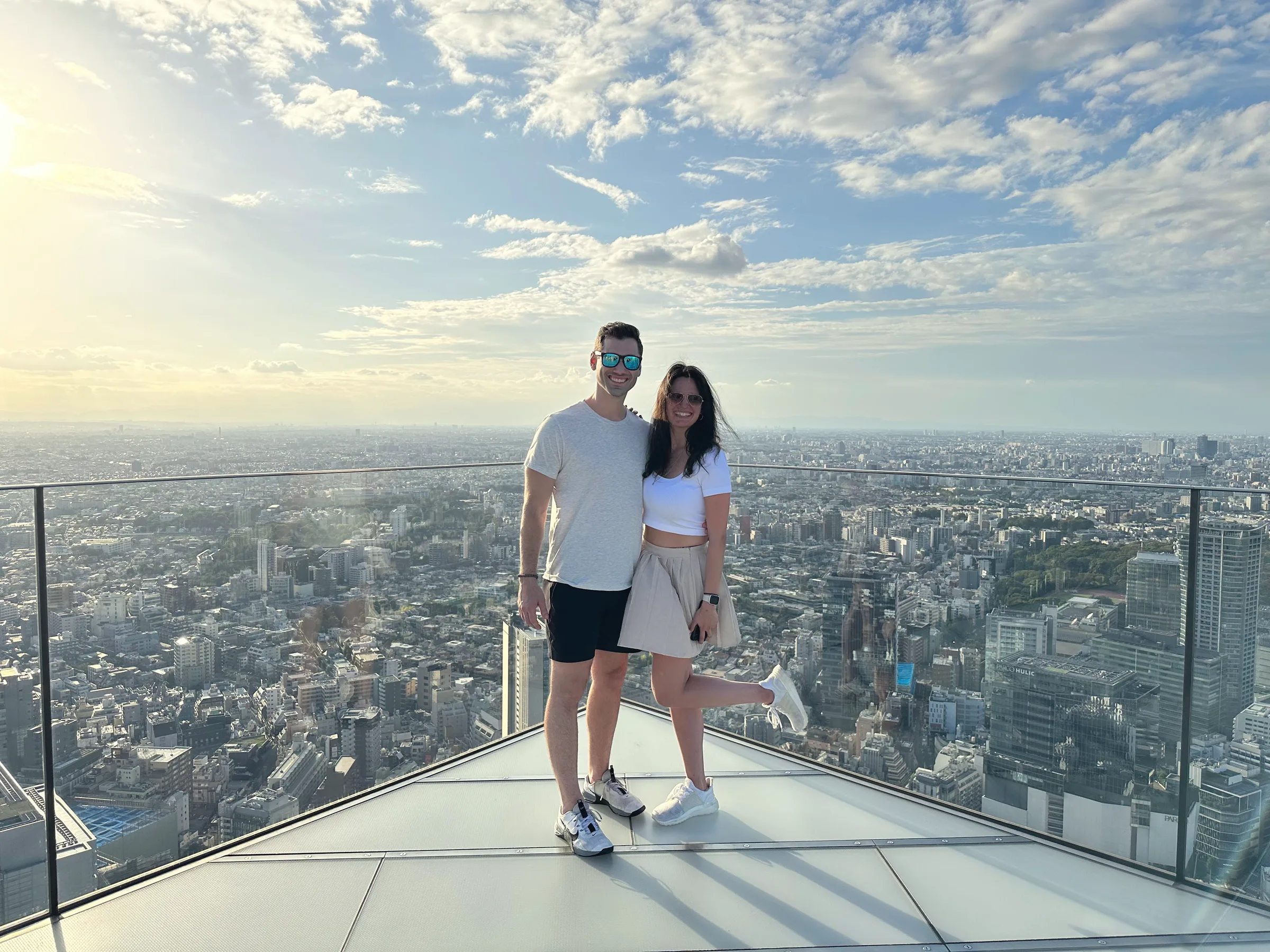 Carmela and Charlie on a glass-floored viewing platform high above a sprawling city under a bright sky