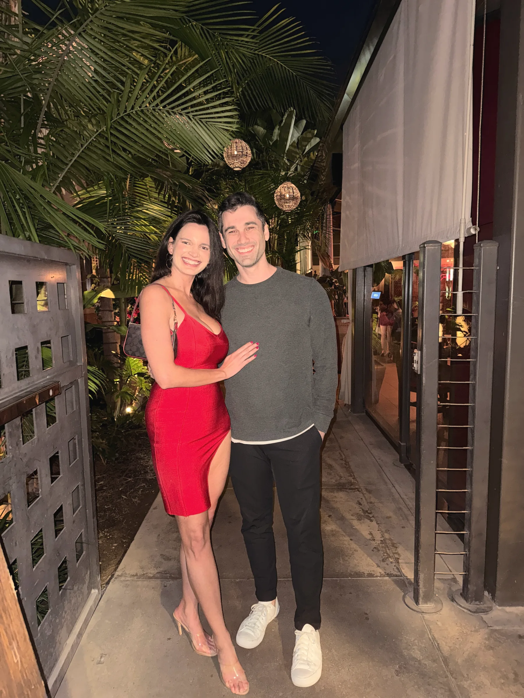 Carmela in a red dress and Charlie in a grey sweater, arms around each other on a path lined with palm leaves and warm lights