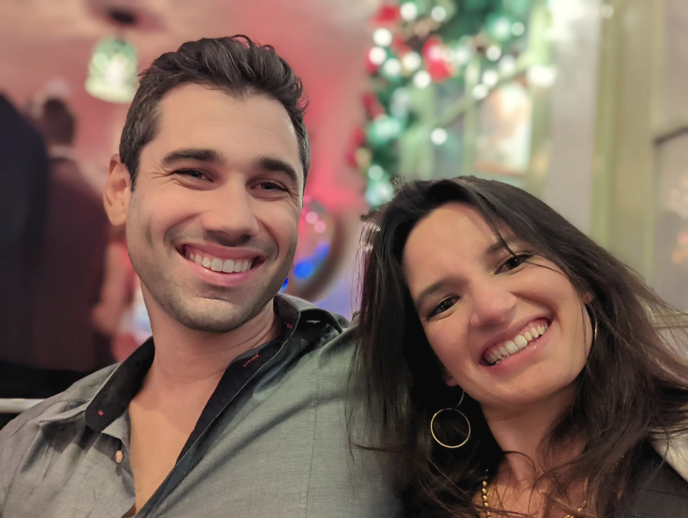 A close selfie of Charlie and Carmela, both beaming, with blurred Christmas decorations behind them