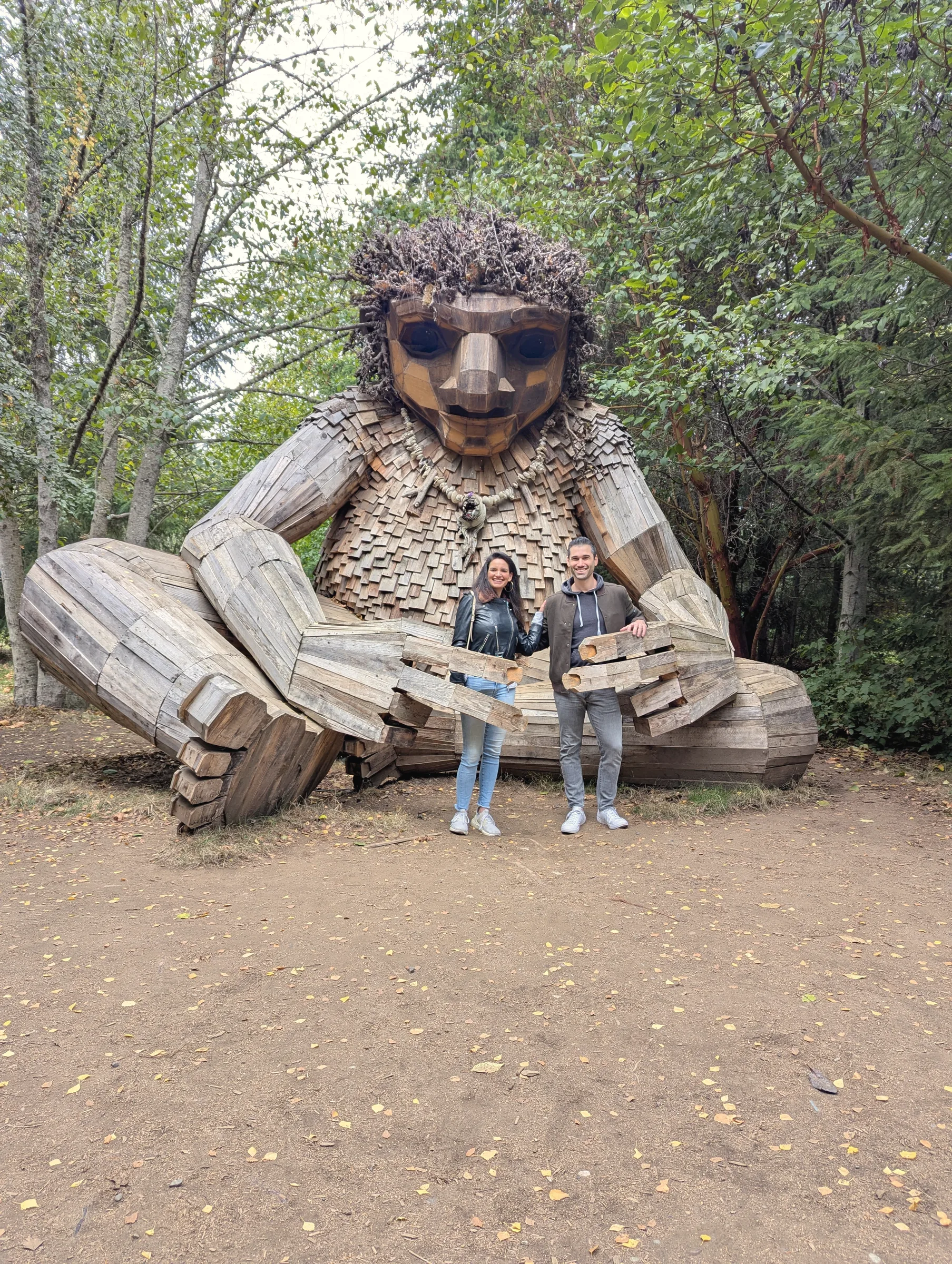 Carmela and Charlie standing in front of a huge seated wooden troll sculpture in a forest clearing
