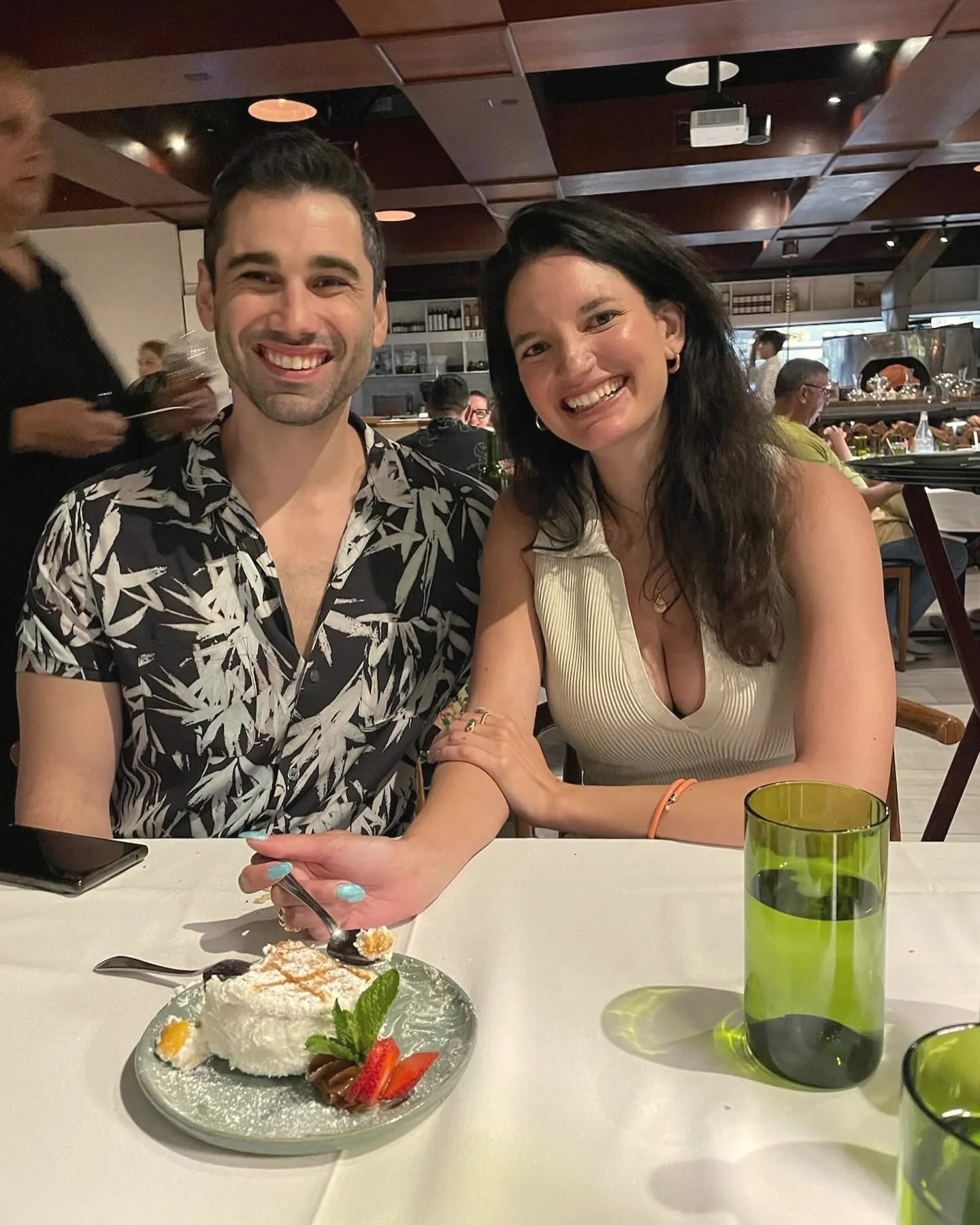 Charlie in a patterned shirt and Carmela smiling at a restaurant table with a plate of dessert in front of them