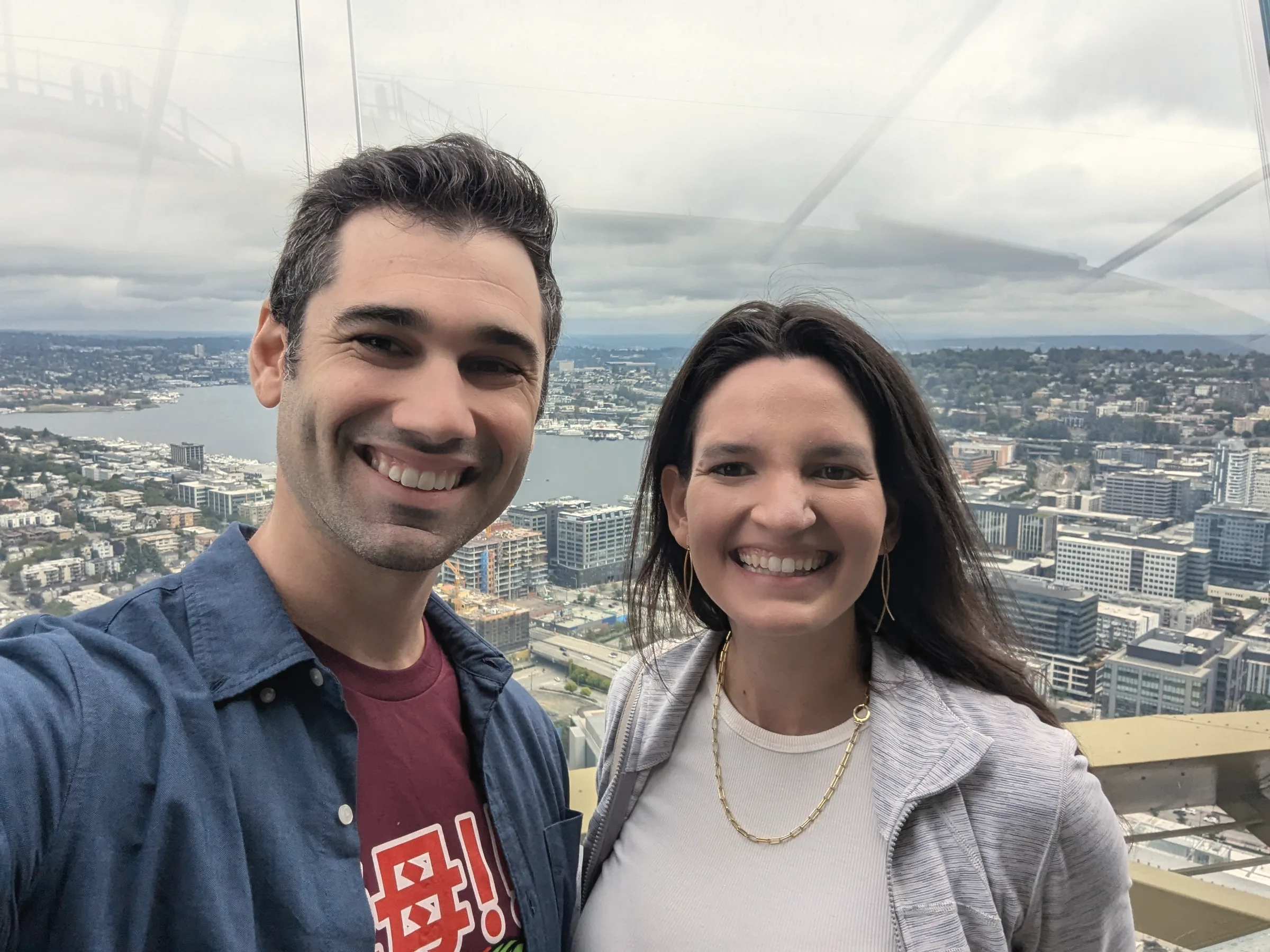 Carmela and Charlie smiling in front of a big window, a harbour and city skyline in the distance