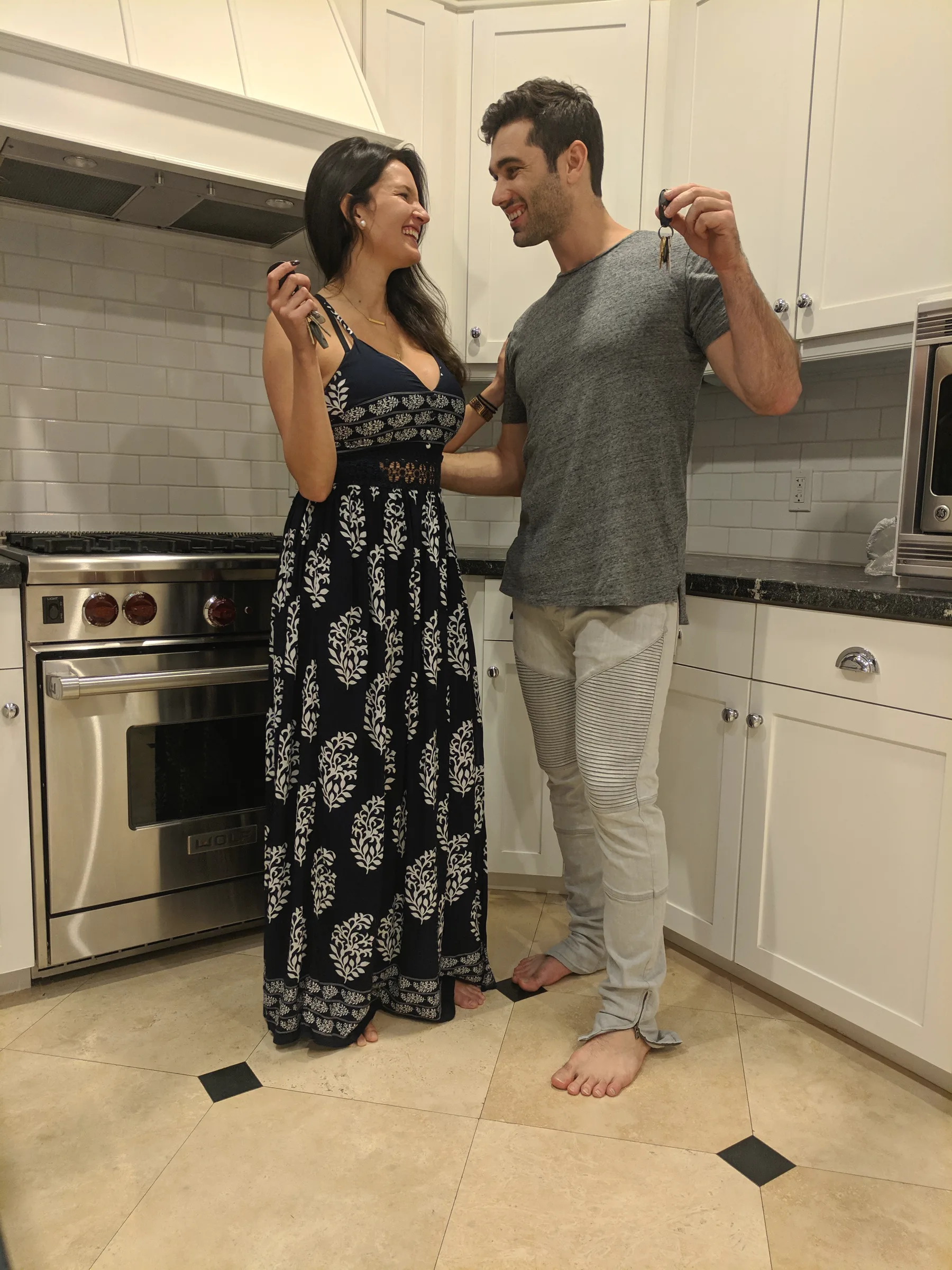 Carmela in a long patterned dress and Charlie in a grey t-shirt, laughing together in a bright white kitchen
