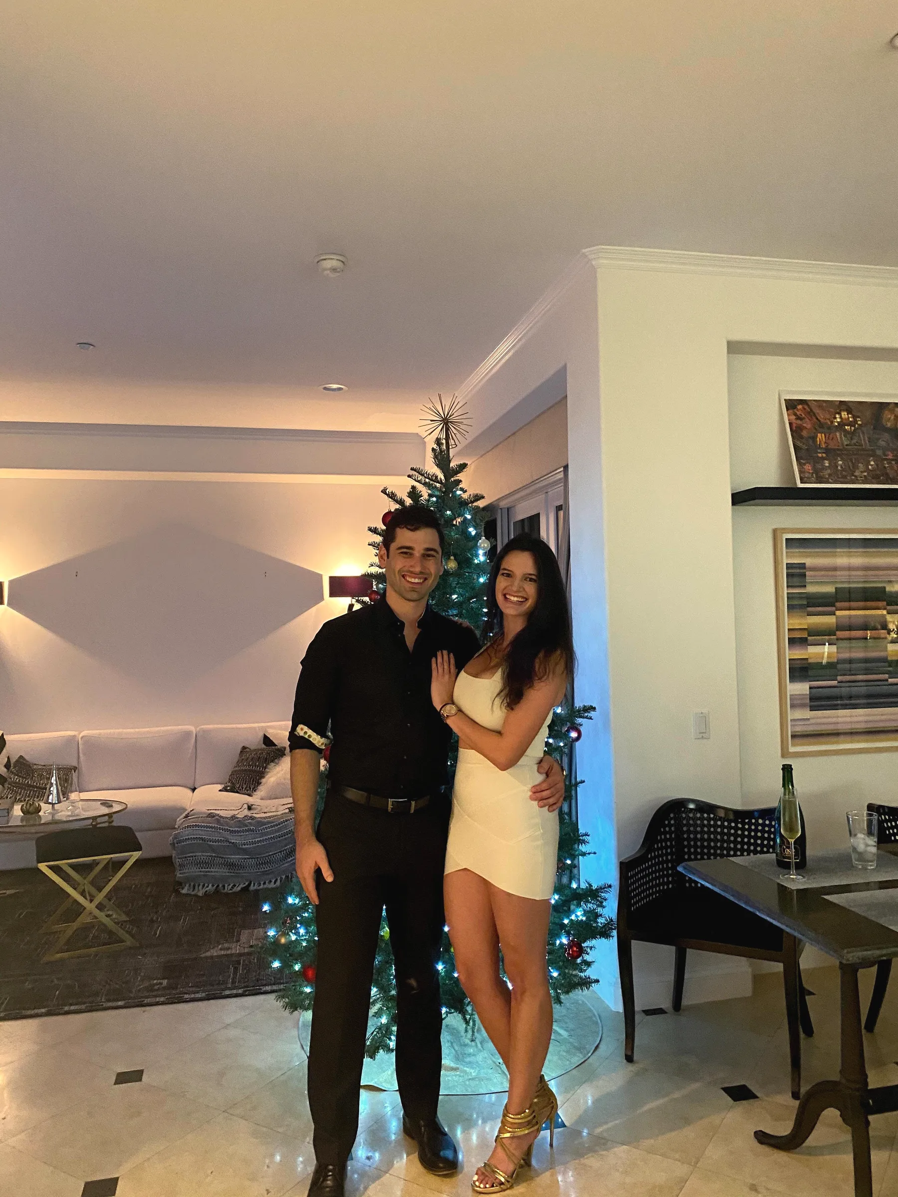 Charlie in black and Carmela in a white dress standing by a decorated Christmas tree in a living room