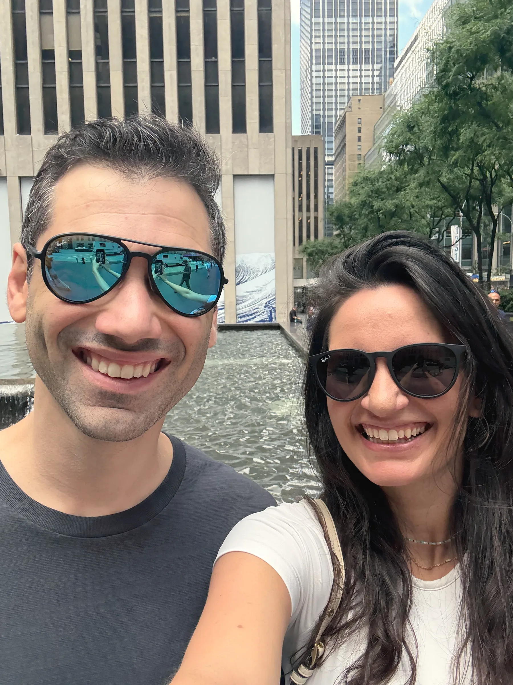 Charlie in mirrored sunglasses and Carmela in dark sunglasses smiling in a selfie in front of tall city buildings and a fountain