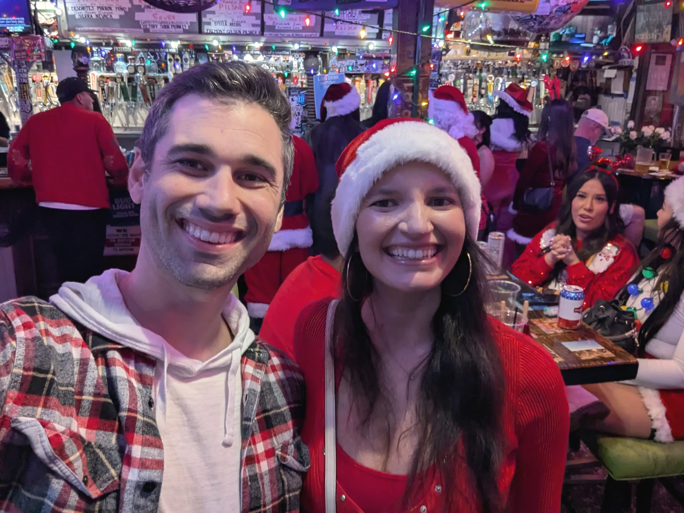 Charlie in a plaid shirt and Carmela in a Santa hat, smiling in a busy bar full of holiday decorations