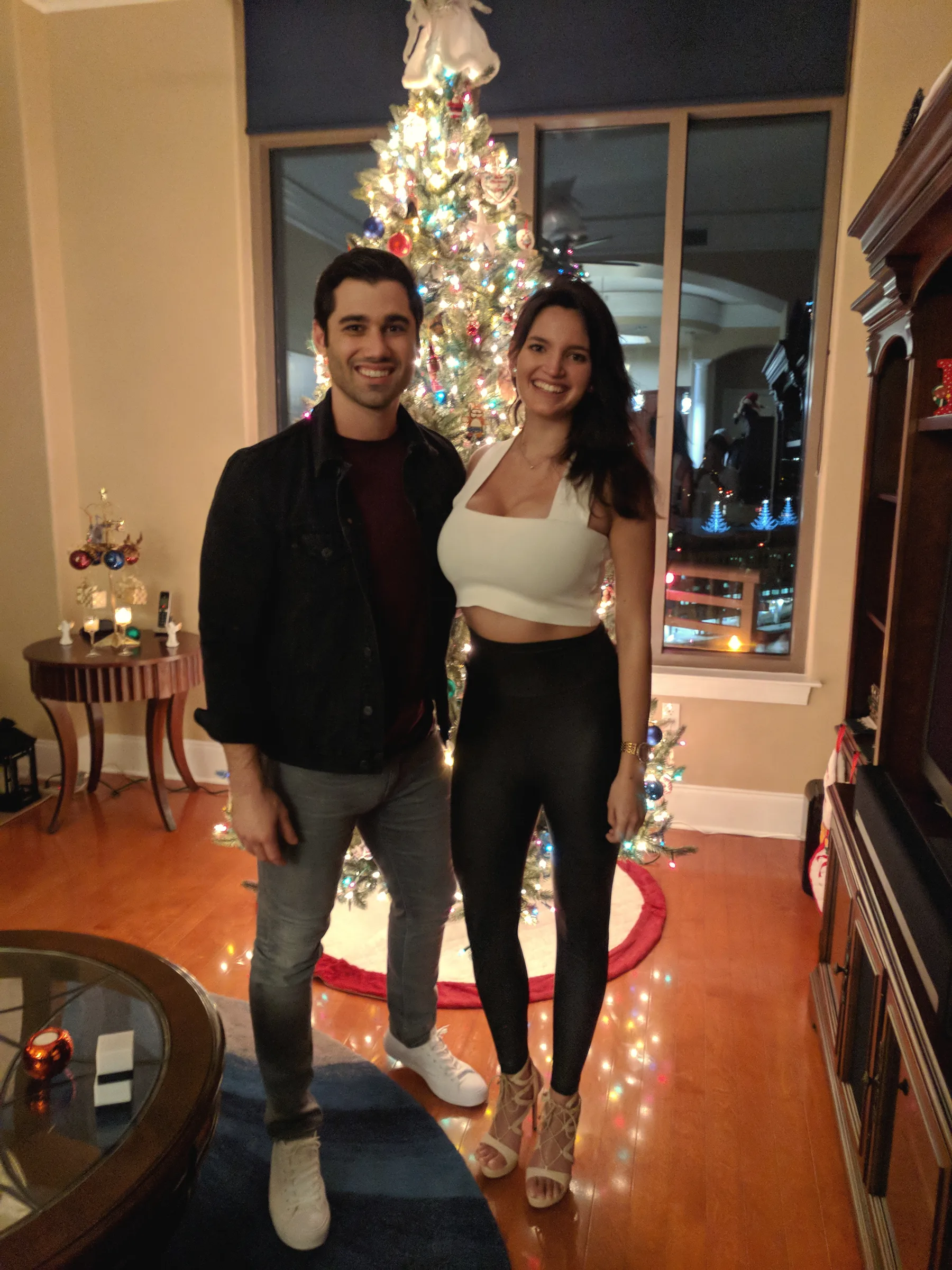 Carmela and Charlie standing by a lit Christmas tree in a living room with a wood floor
