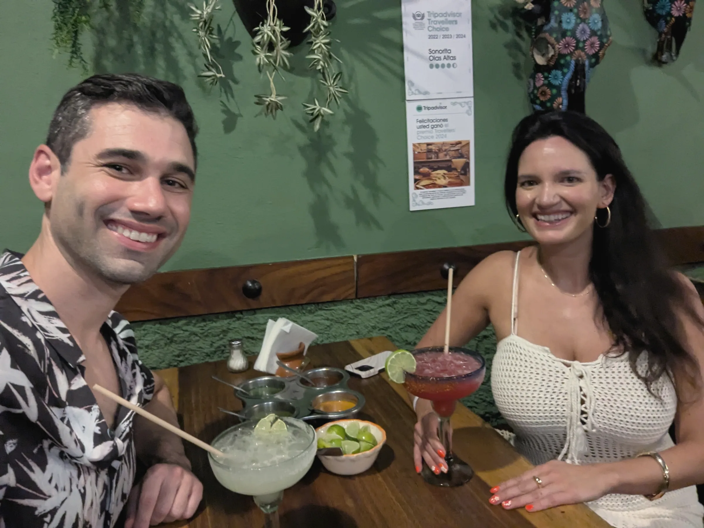 Charlie in a palm-print shirt and Carmela in a white top at a restaurant table with a large margarita and a bowl of limes