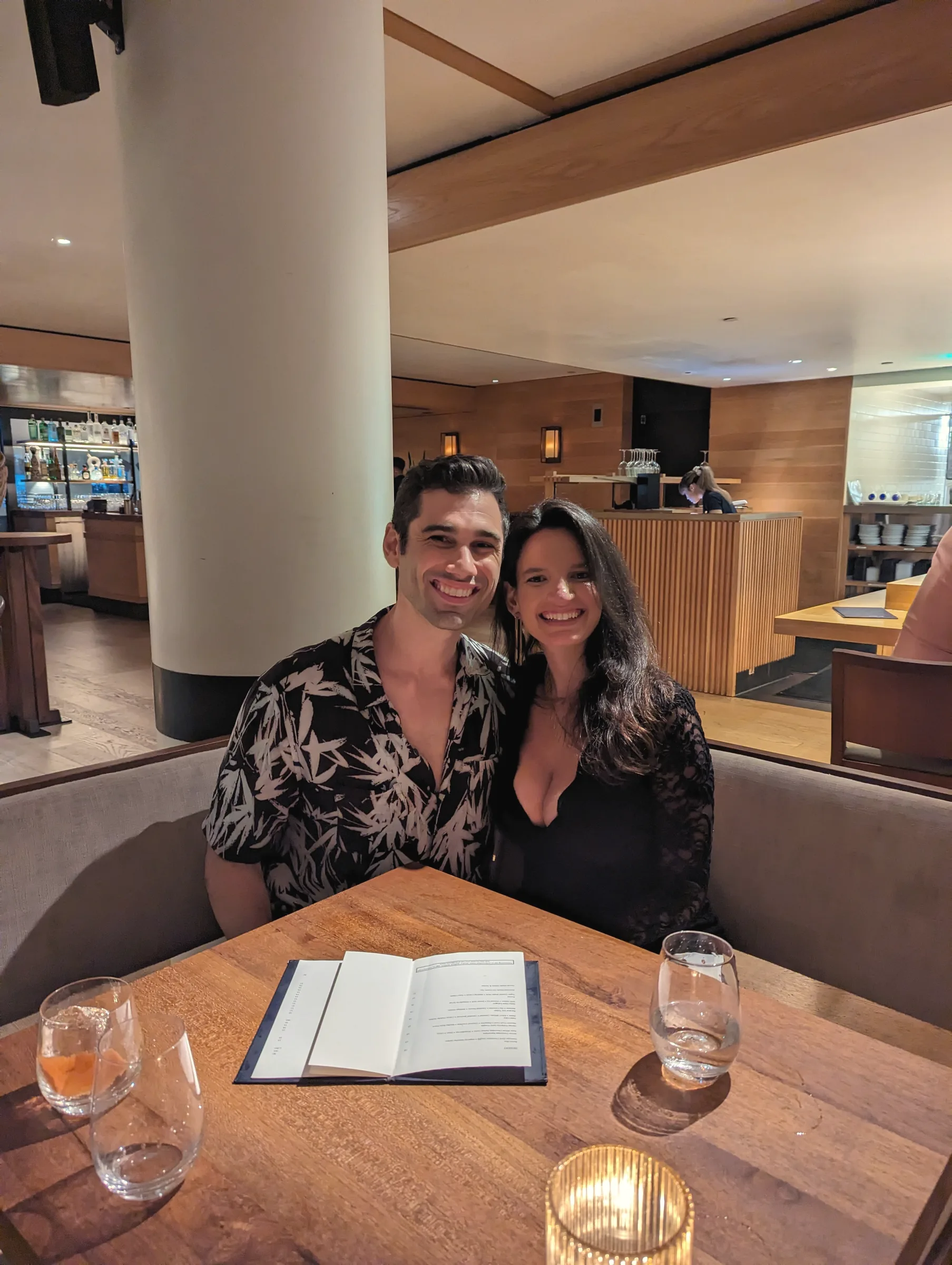 Charlie and Carmela side by side in a restaurant booth with drinks and a menu on the table