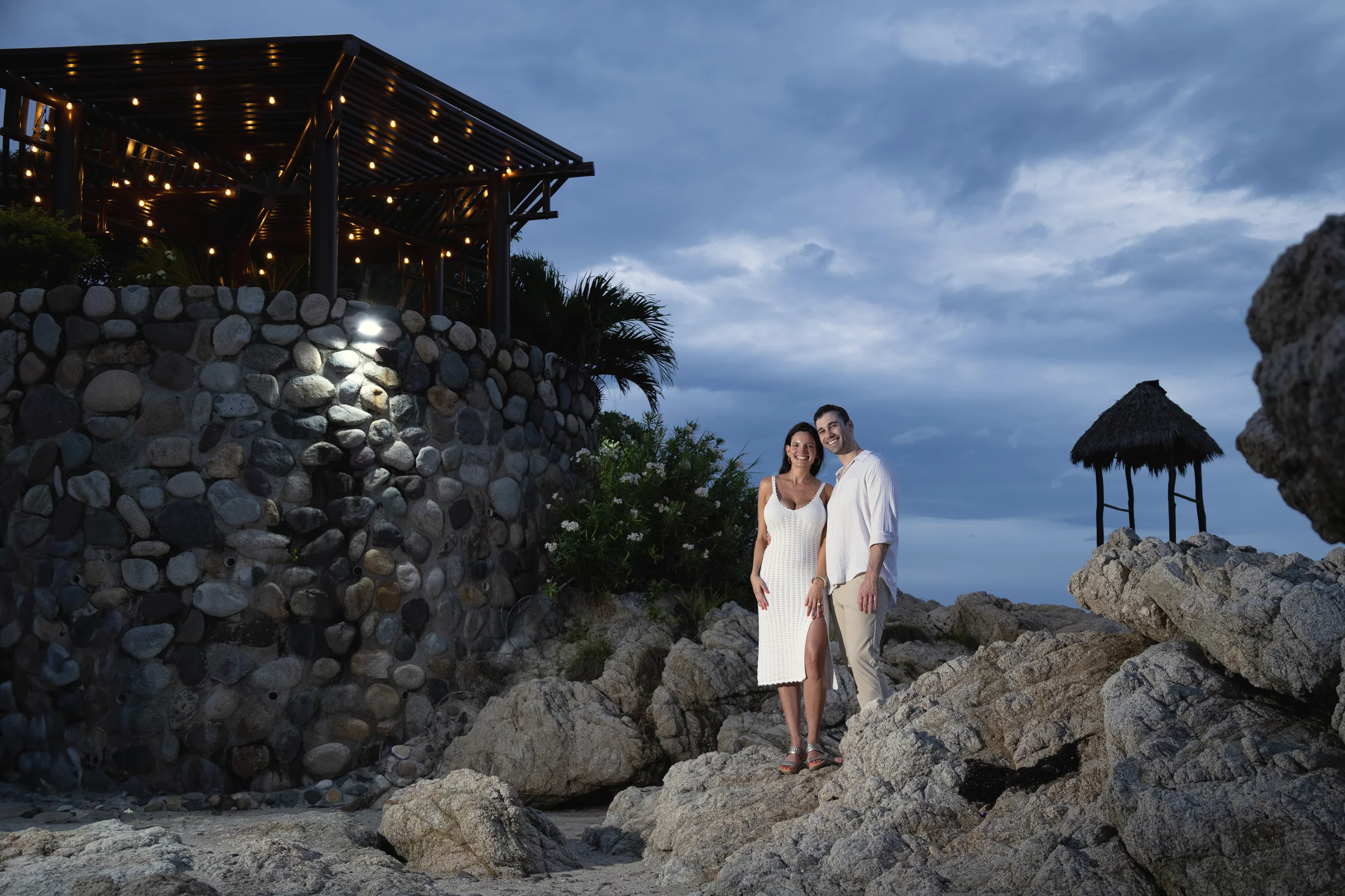 Carmela and Charlie small in the frame on a rocky shore at dusk, a stone wall and a palapa above them and a stormy blue sky