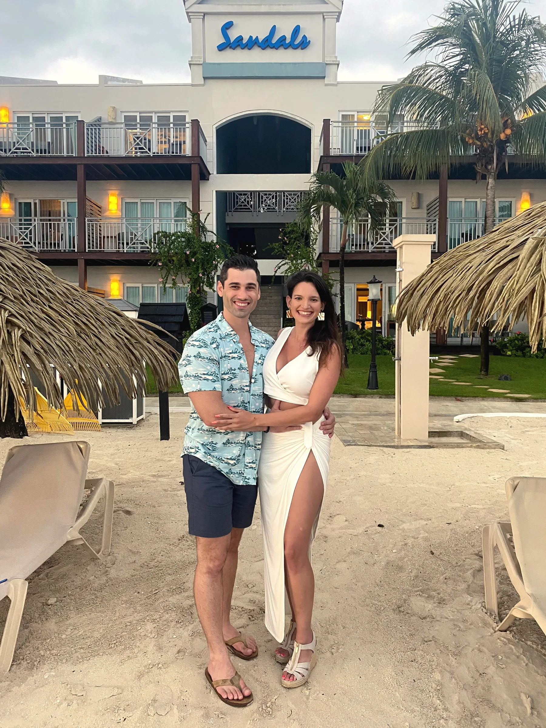 Carmela in a white dress and Charlie in a floral shirt on a sandy beach in front of a resort building with thatched umbrellas
