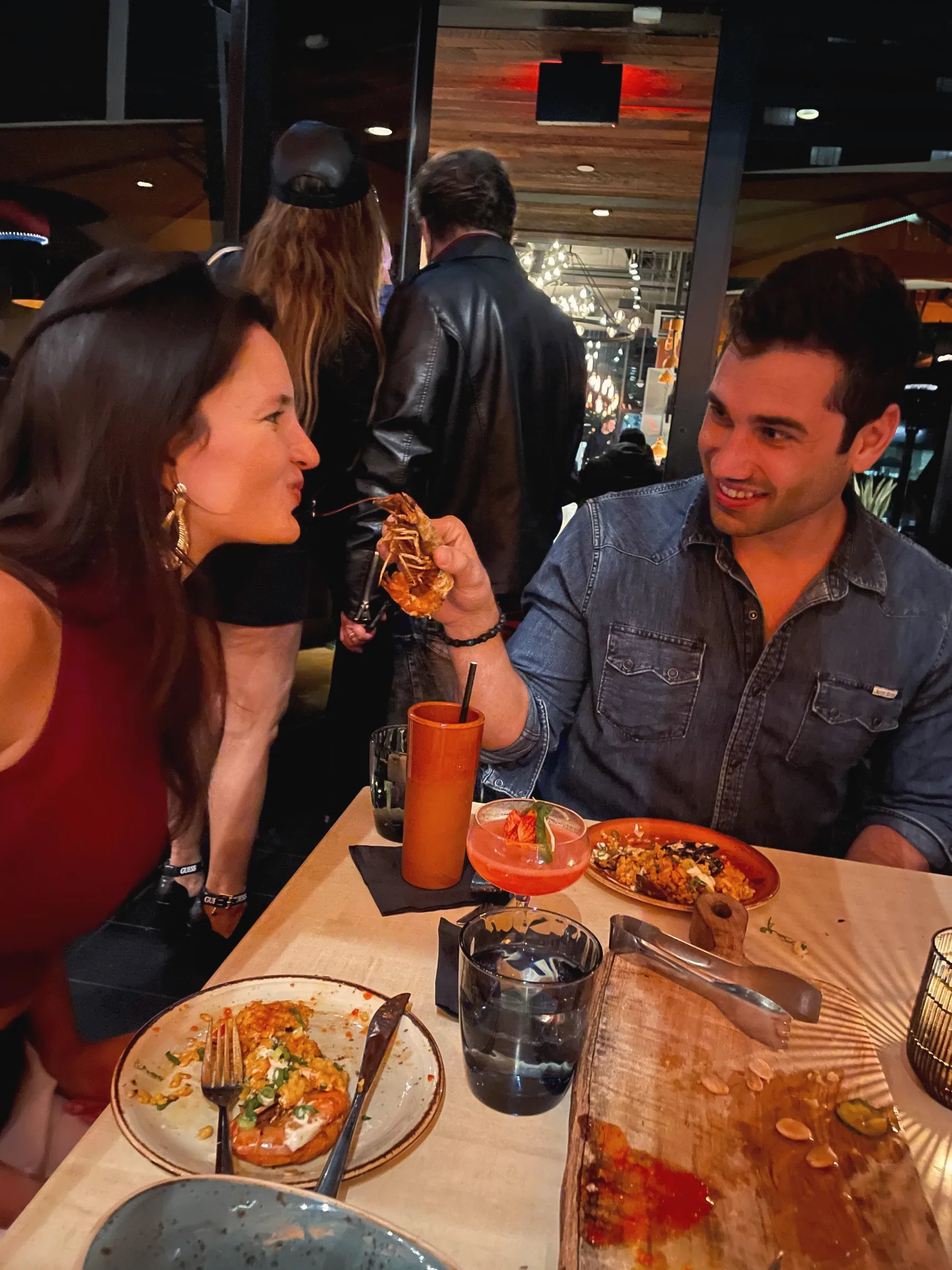 Charlie offering Carmela a bite from his fork across a crowded restaurant table with plates and drinks