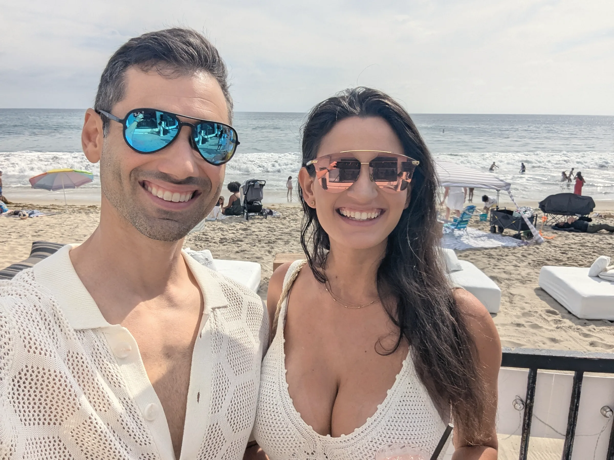 Carmela and Charlie in sunglasses smiling together, a sandy beach with umbrellas behind them