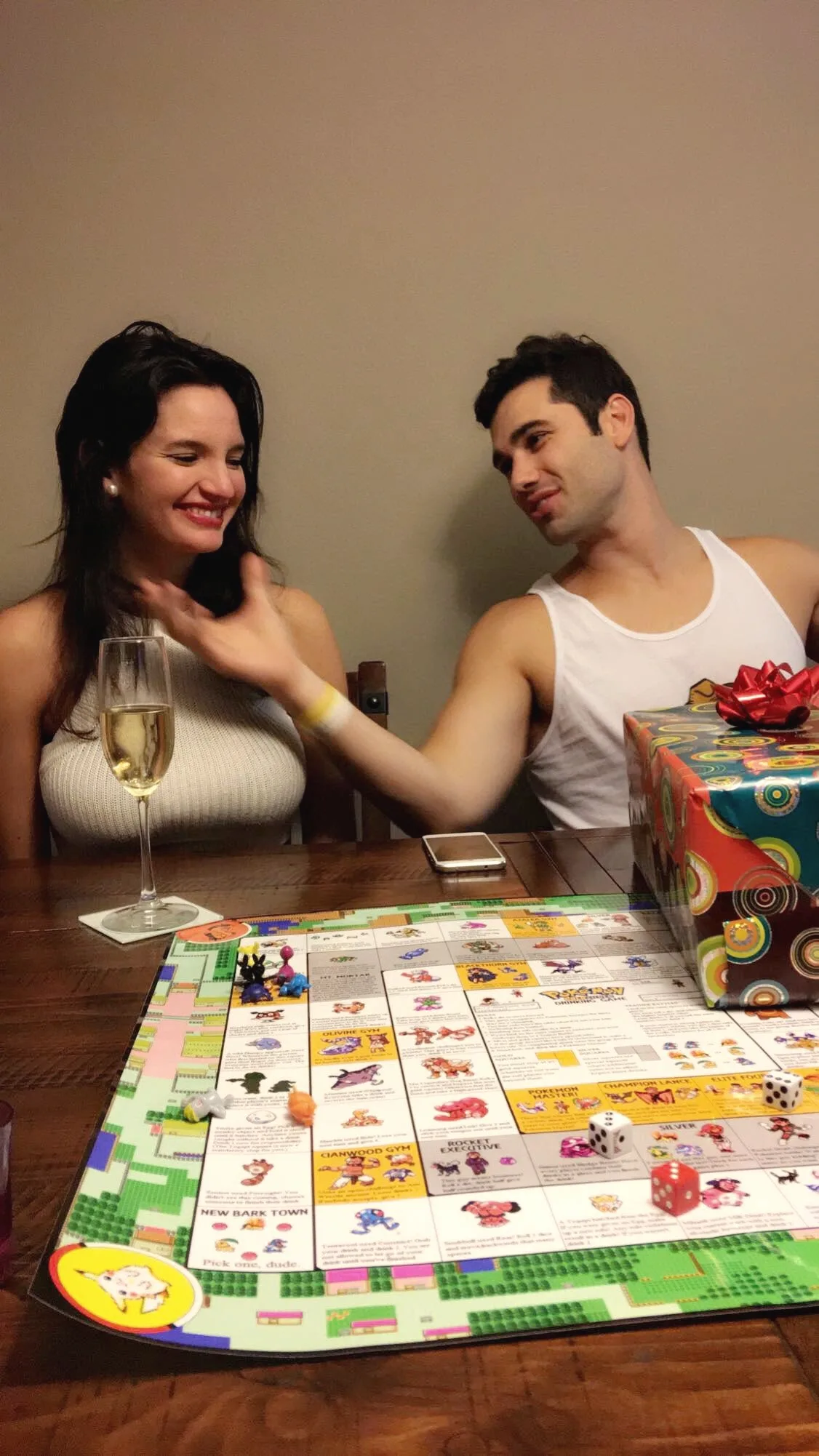 Carmela laughing with a glass of champagne while Charlie leans in beside a colourful board game on a wooden table