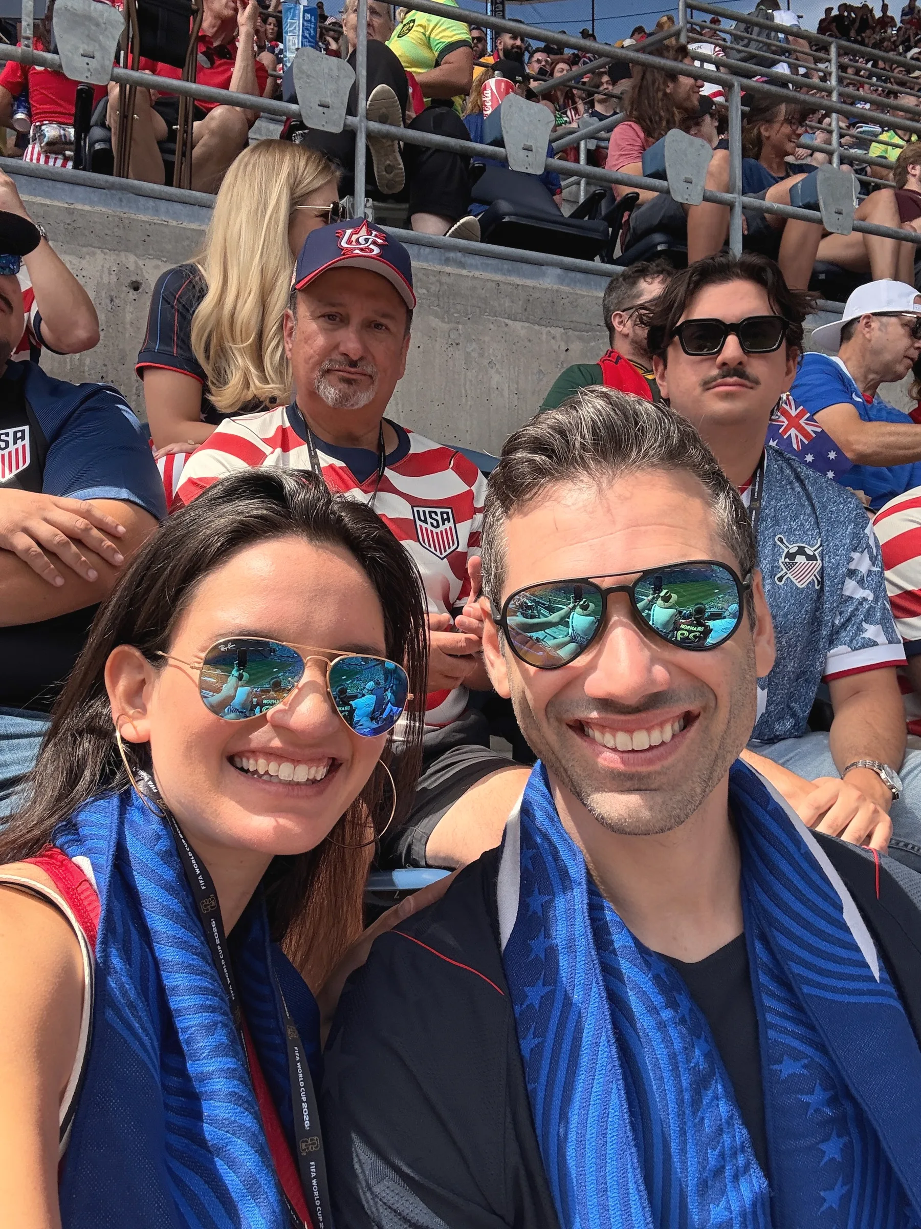 Carmela and Charlie in sunglasses and matching blue scarves, smiling in a crowded stadium stand