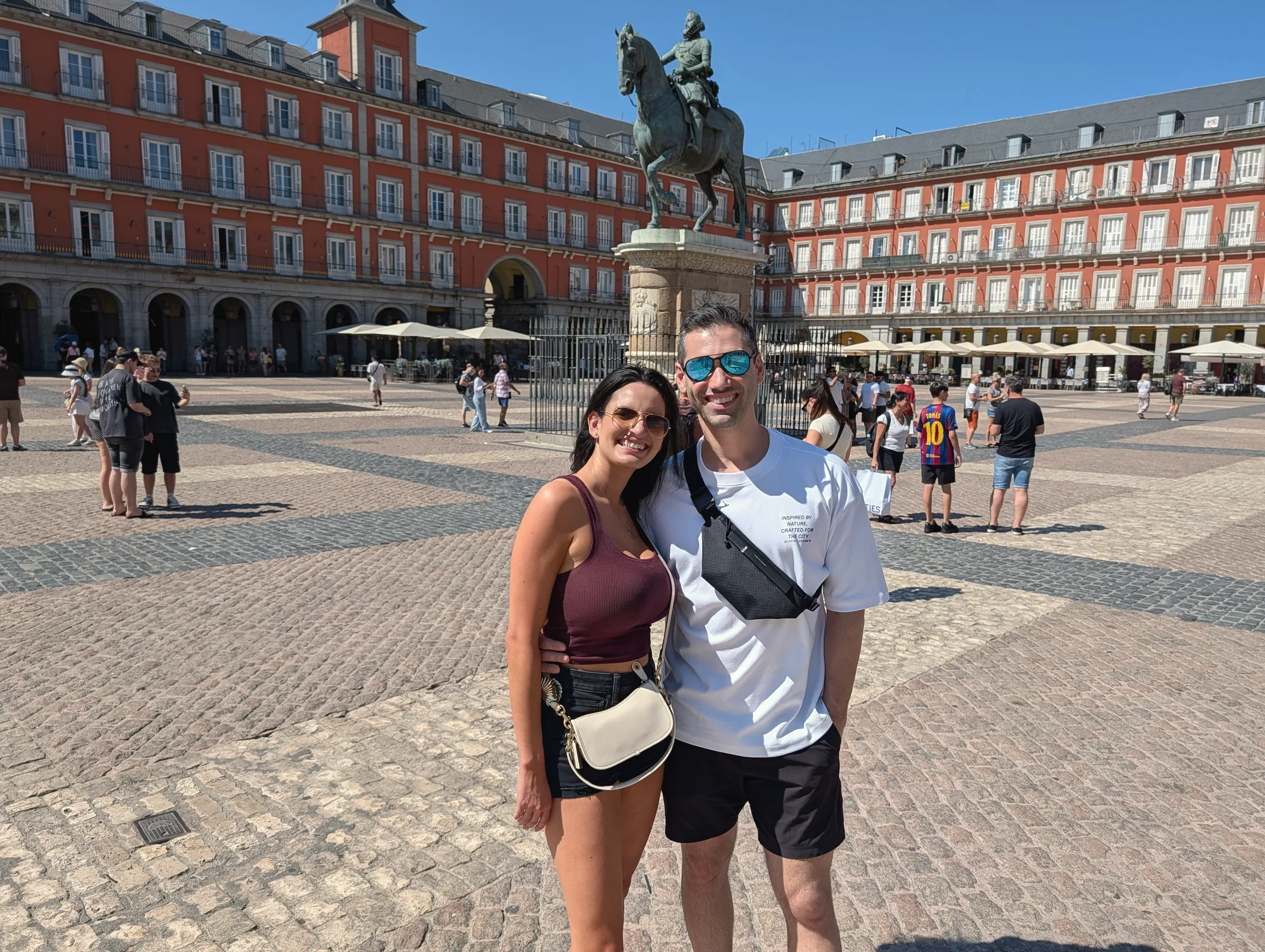 Carmela and Charlie posing in a large cobbled square with red-brick arcaded buildings and a statue on a horse behind them