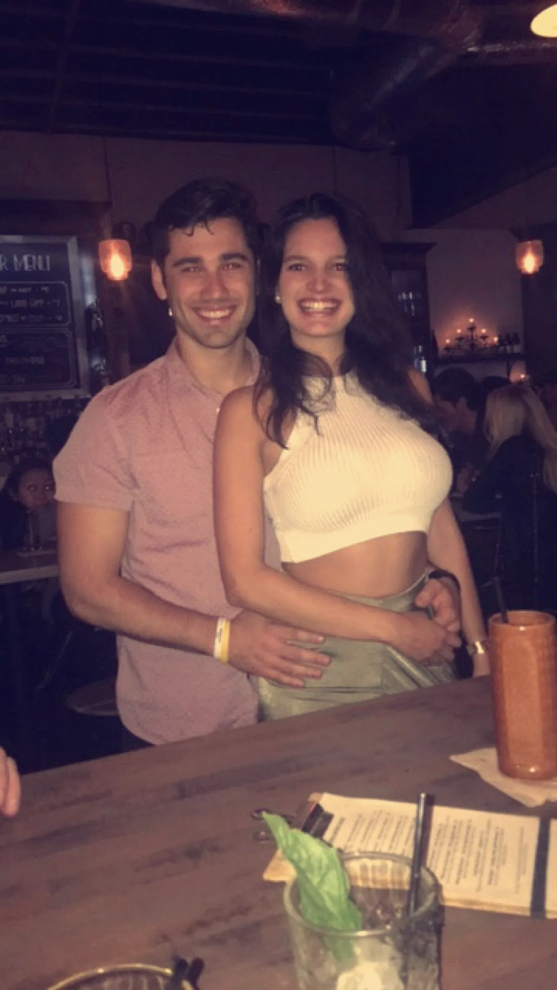Carmela and Charlie seated close at a wooden table in a dim restaurant, a cocktail in a copper mug in front of them