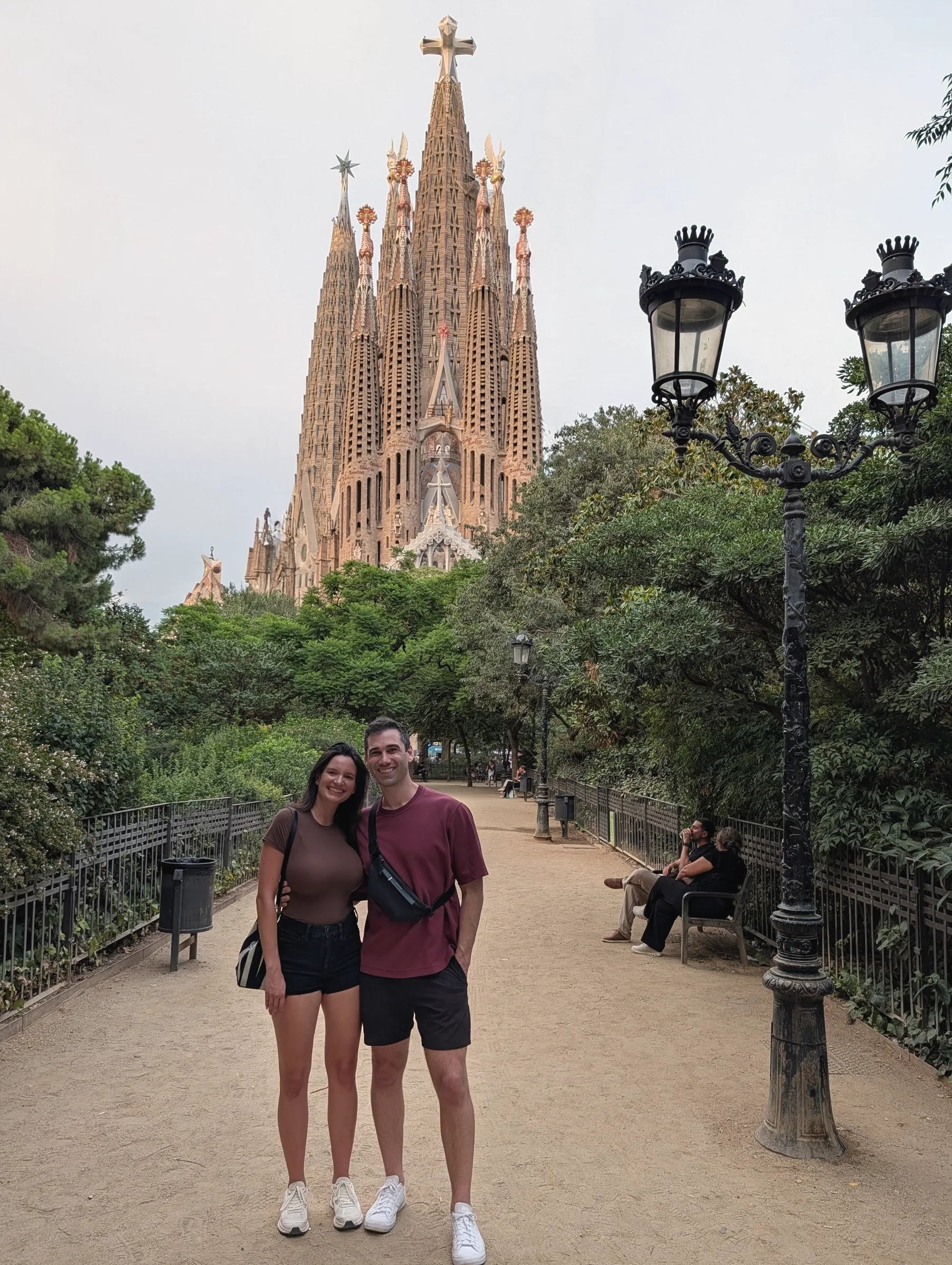 Carmela and Charlie small on a sandy path lined with lamp posts, a tall ornate church with spires rising over the trees