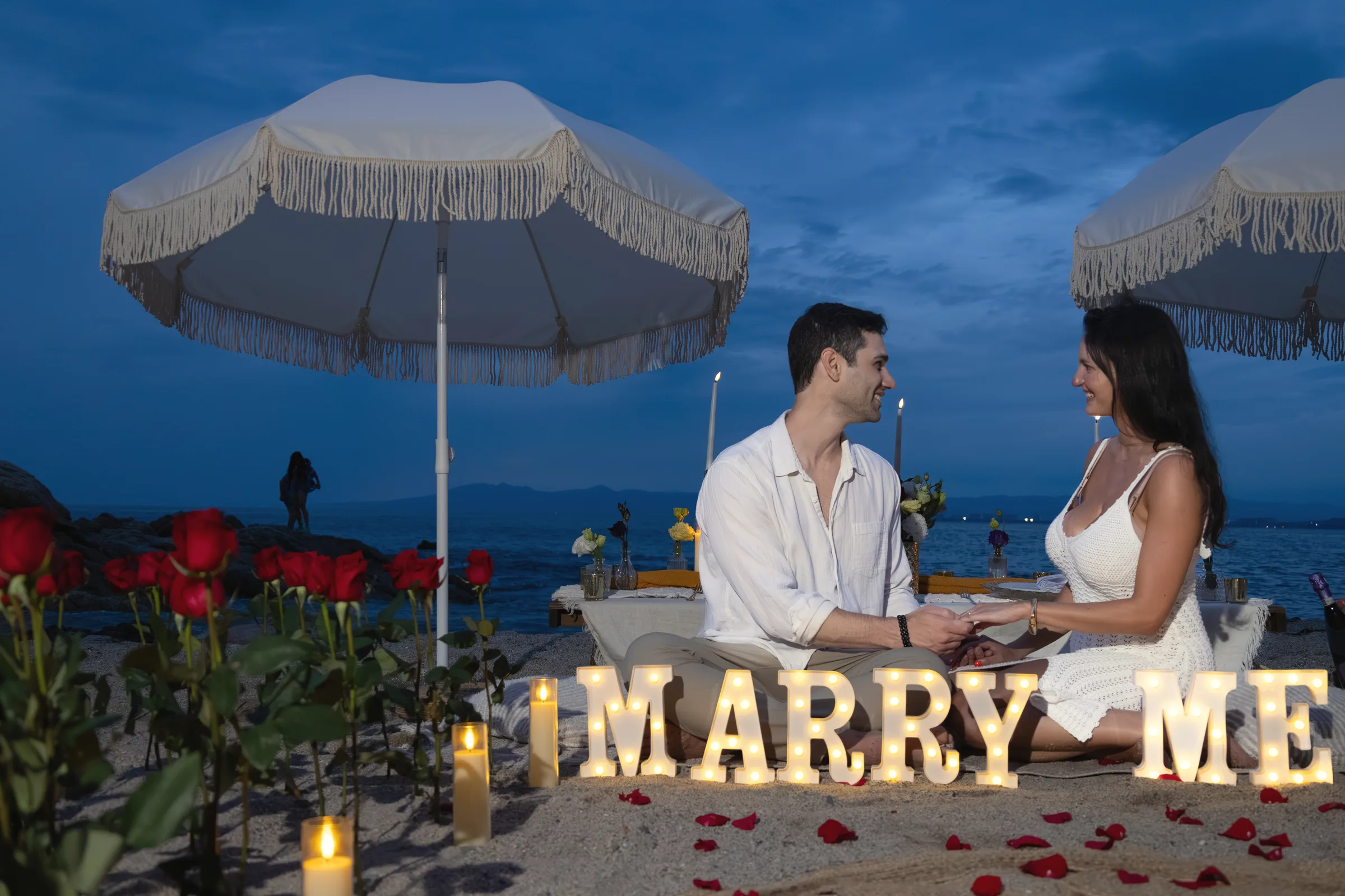 Carmela and Charlie sitting together holding hands beside a glowing MARRY ME sign, roses and a white umbrella around them at dusk