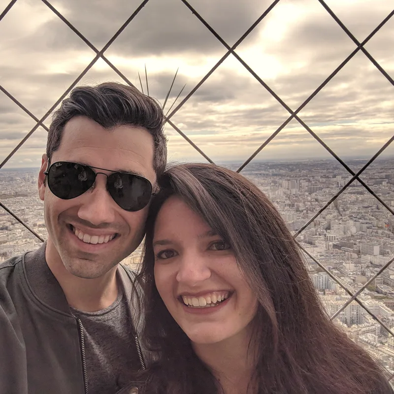 Carmela and Charlie smiling in a selfie high up in the Eiffel Tower, Paris spread out behind them