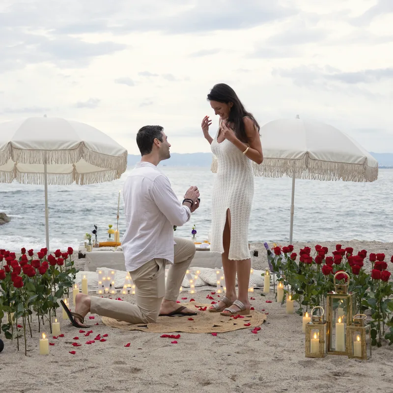 Charlie down on one knee proposing to Carmela on a beach, with cream parasols, red roses and candles around them