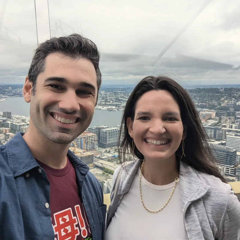 Carmela and Charlie smiling in a selfie high above a waterfront city, with the skyline behind them